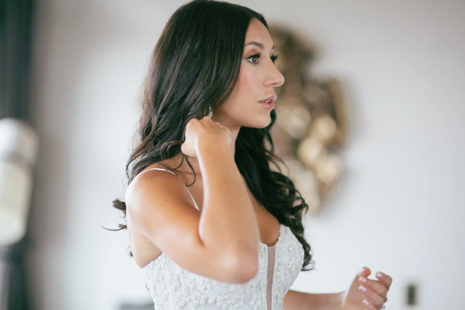 A woman with long dark hair, wearing a white dress, standing indoors, adjusting her earring.