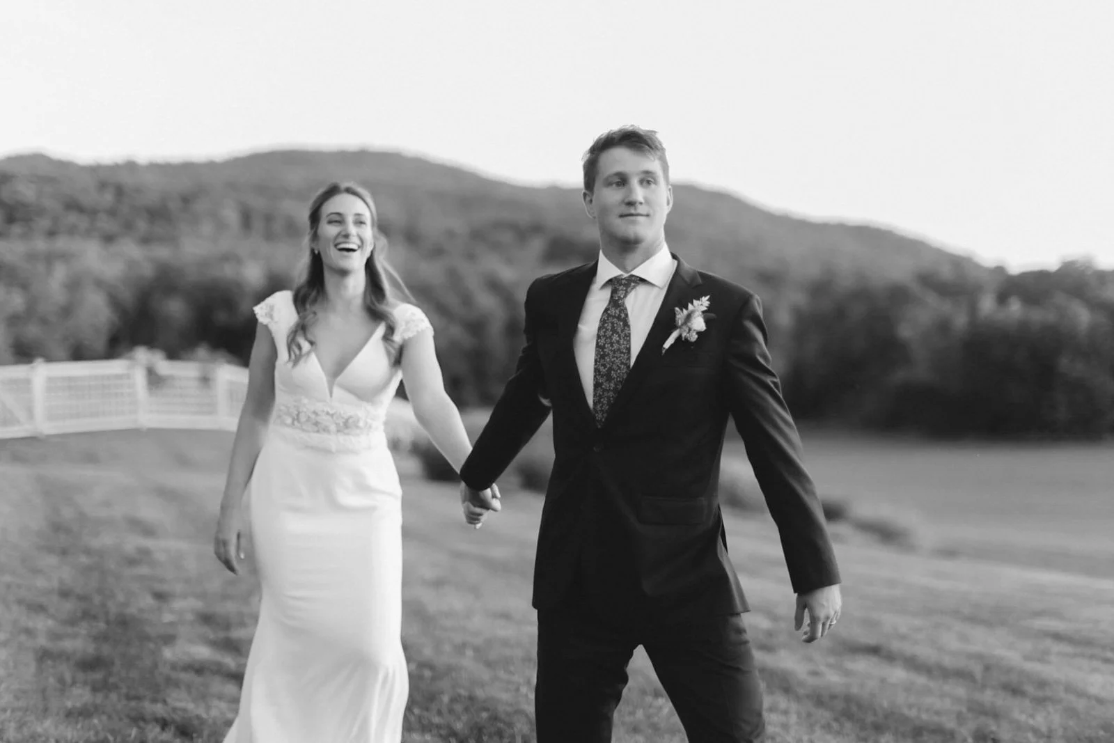 A newlywed couple holding hands outdoors, walking on a grassy field with hills and a fence in the background. The bride is laughing, wearing a white wedding dress, and the groom is dressed in a dark tuxedo with a boutonnière.