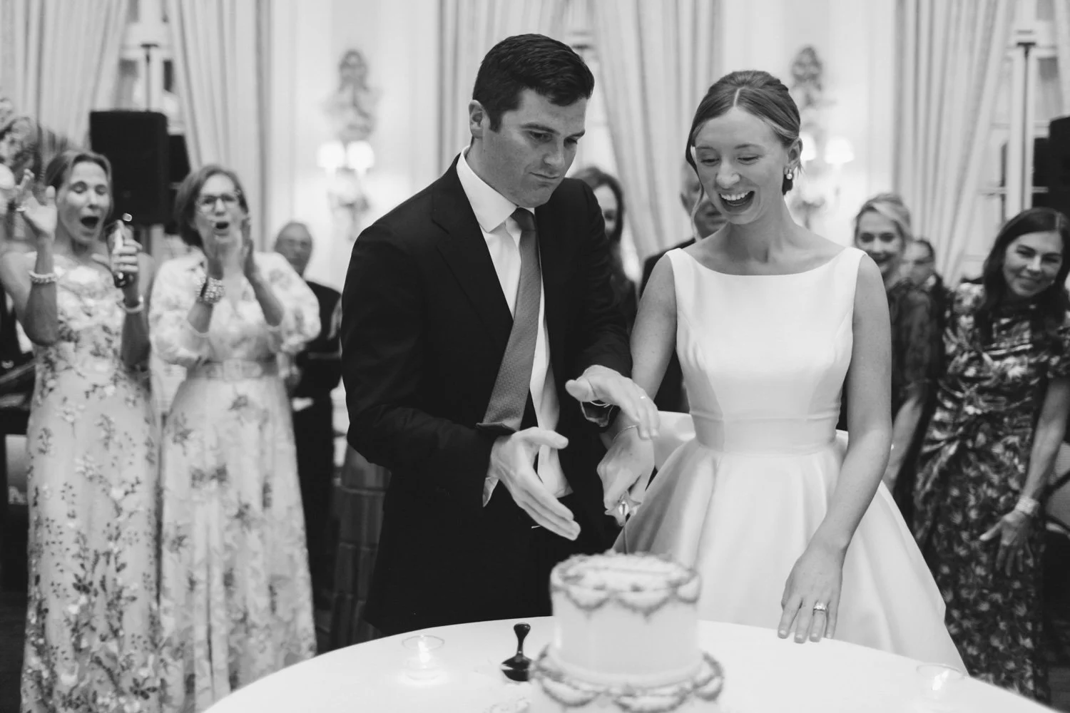 A bride and groom cutting a wedding cake at their reception, surrounded by happy guests.