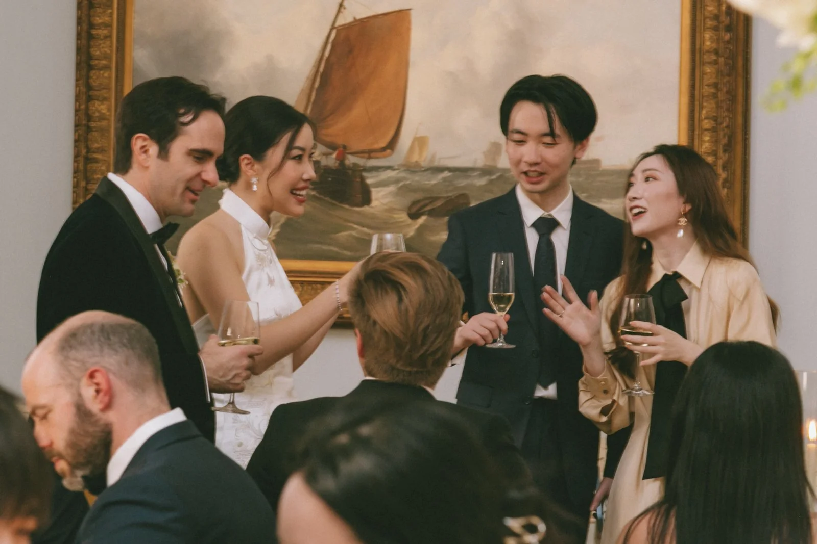 People celebrating at a formal dinner party, smiling and holding glasses of champagne, with a ship painting hanging on the wall behind them.
