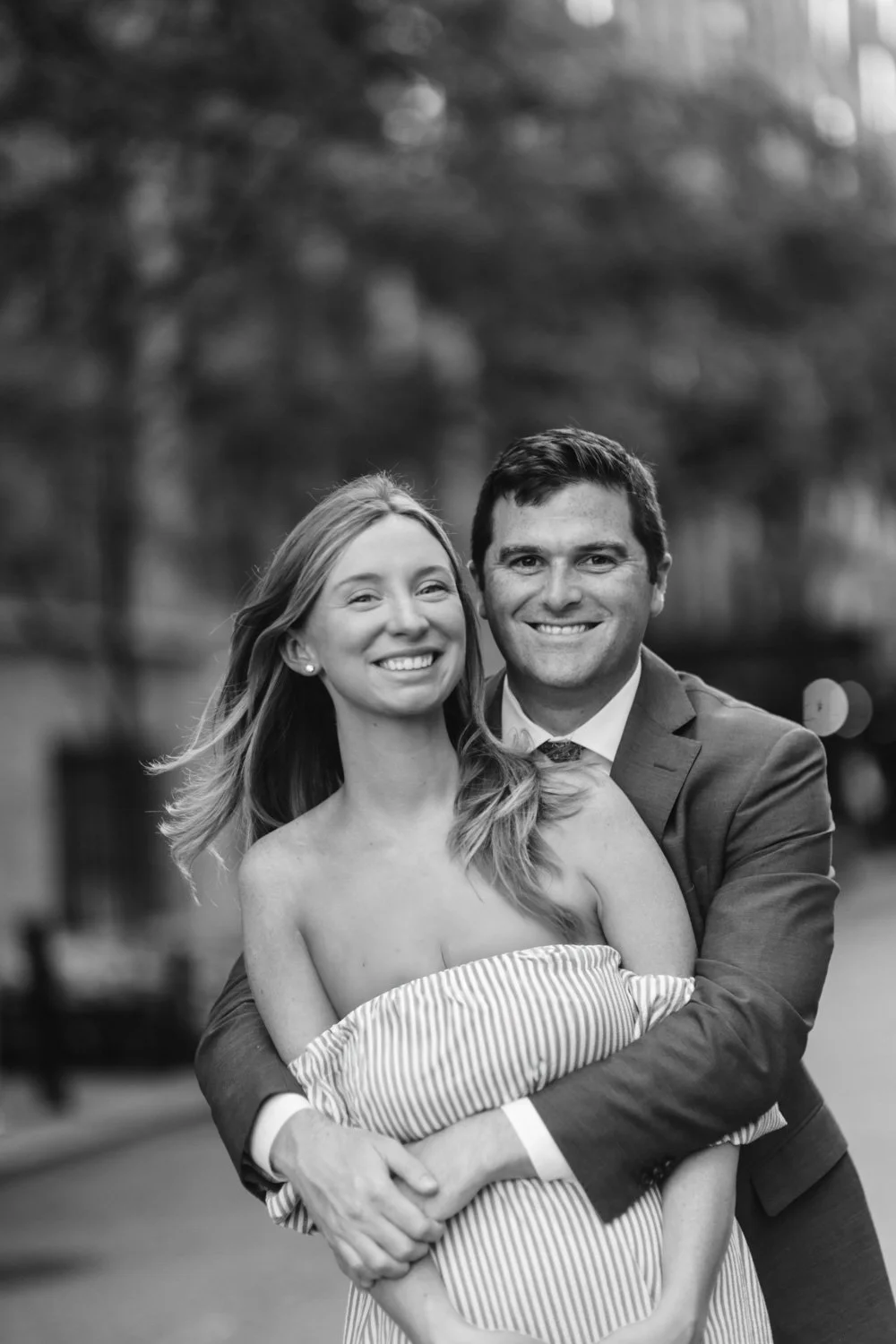 A smiling couple embracing outdoors, the woman wearing a strapless dress, and the man in a suit, black and white photo.