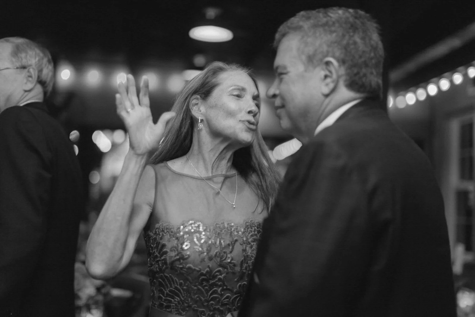 A woman and a man engaged in a close conversation at a social event, with blurred background lights and other attendees visible.