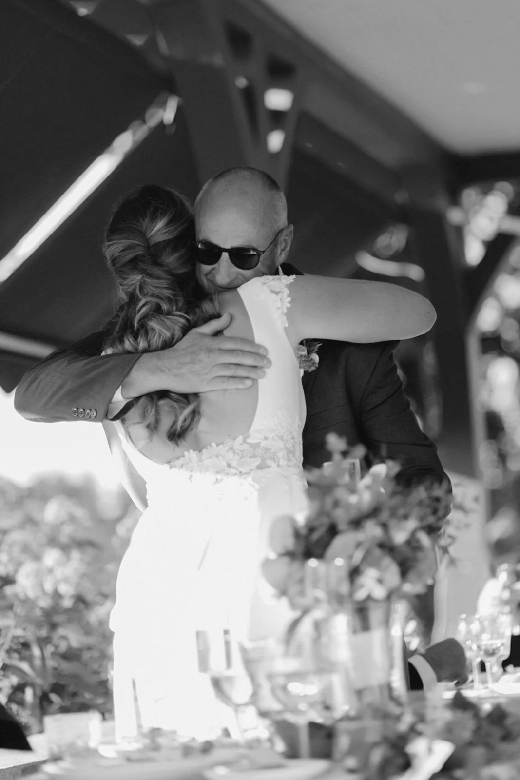 A black and white photo of a man and woman embracing at a wedding. The man is wearing sunglasses and a suit, and the woman is in a wedding dress. They are sharing a heartfelt hug at a celebration table with flowers and drinks.
