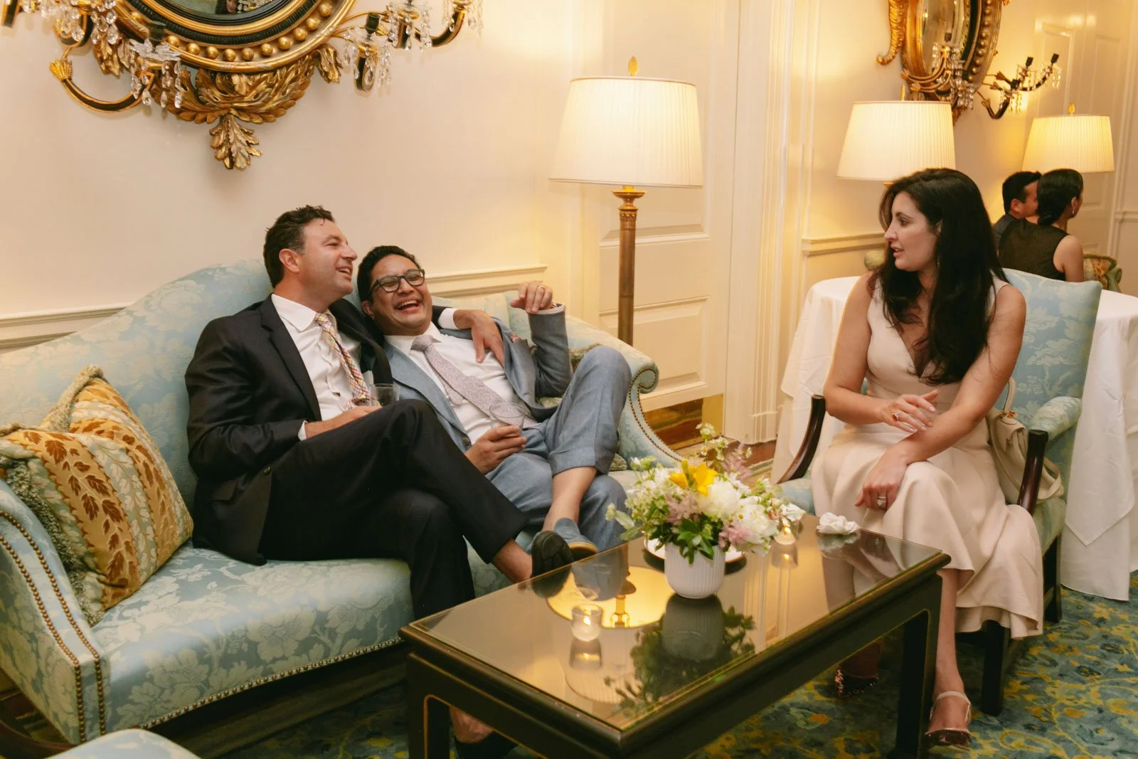 Three people seated in a fancy room, two men in suits laughing and one woman in a cream dress smiling and looking at the men, with a coffee table, flower arrangement, and lamps in the background.