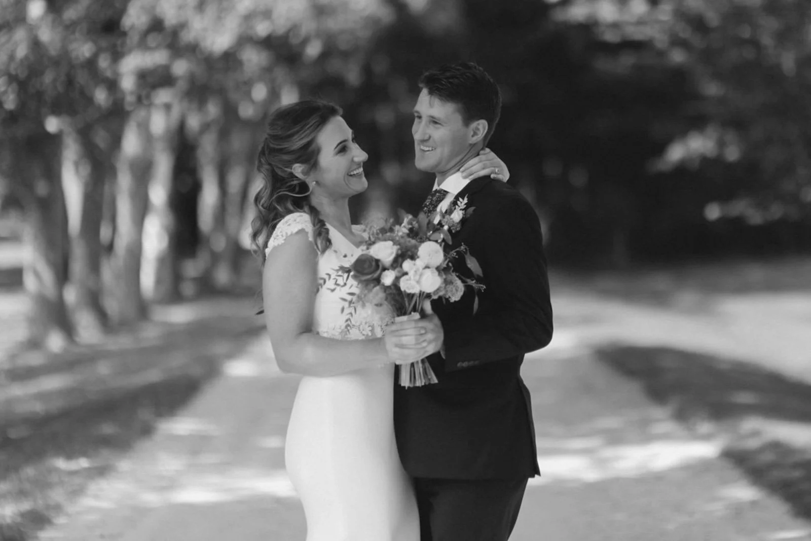A black and white photo of a bride and groom smiling and embracing outside, with trees in the background. The bride is holding a bouquet of flowers.
