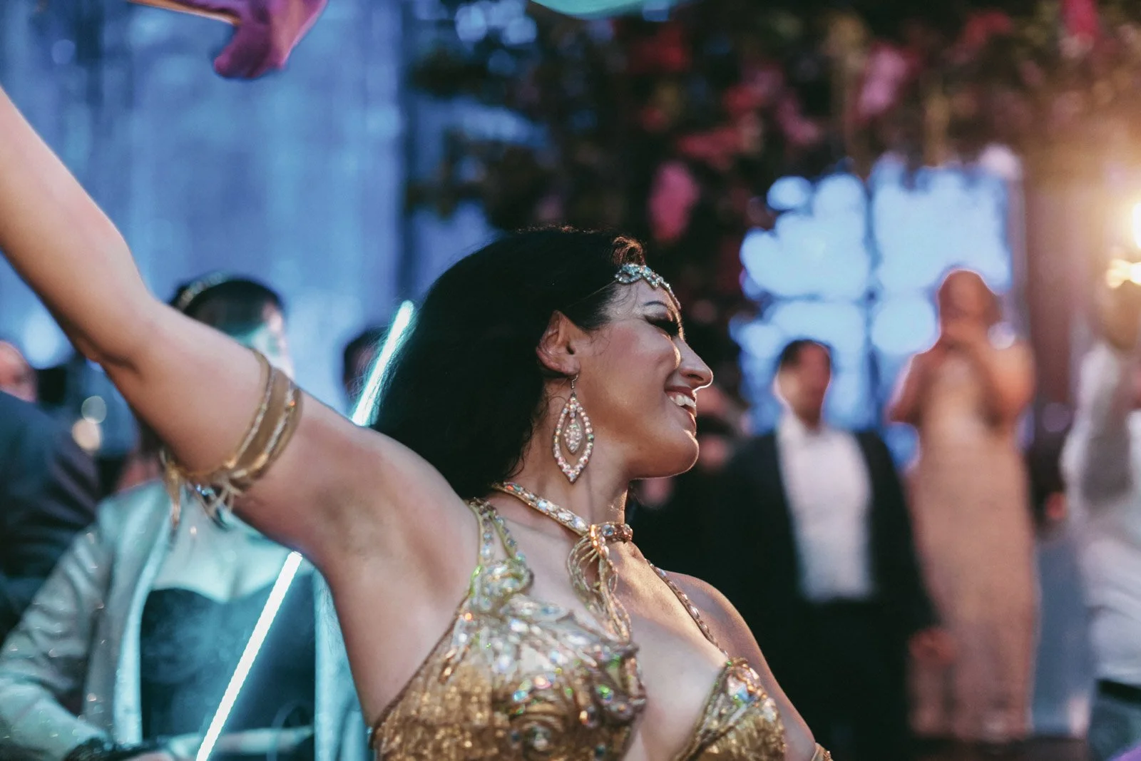 A woman dressed in glamorous gold attire with jewelry, dancing happily at an event or celebration, with other people blurred in the background.
