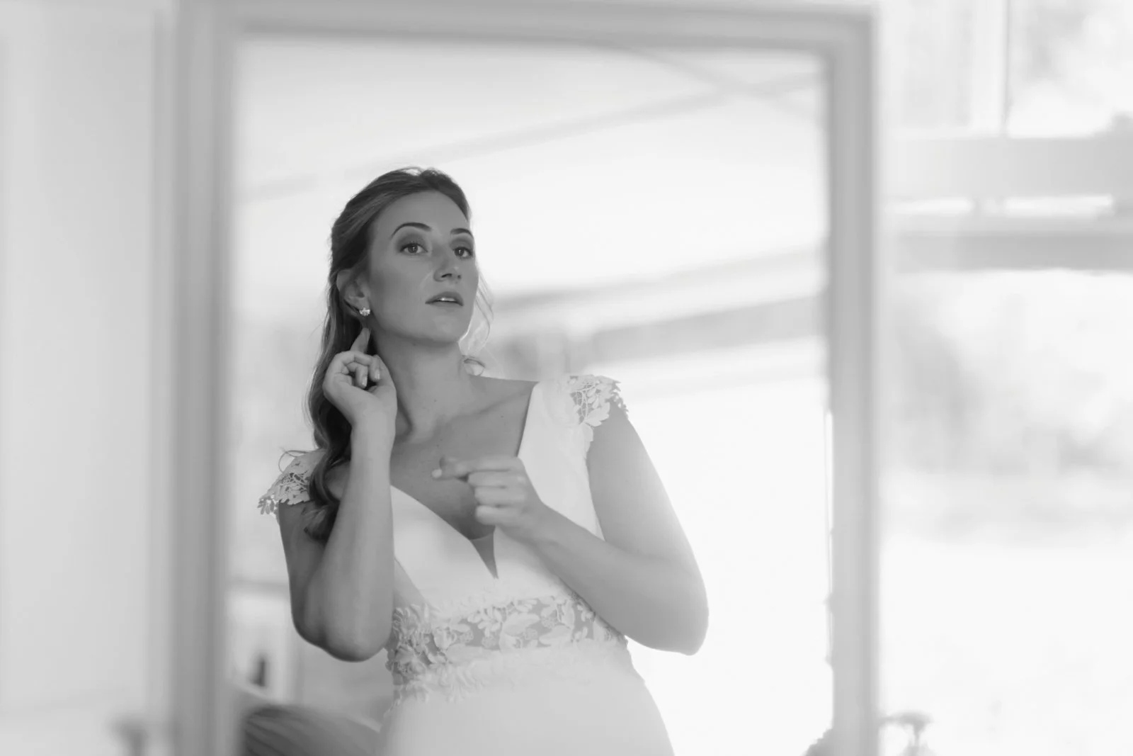 A woman in a wedding dress looking into a mirror, adjusting her earring.