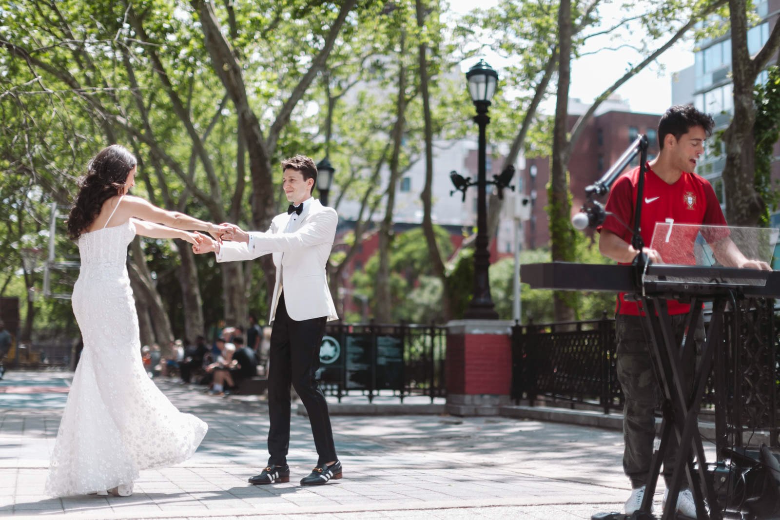 A couple dancing in a park, with a man in a white tuxedo and a woman in a white dress, while a musician in a red sports jersey plays keyboard nearby.