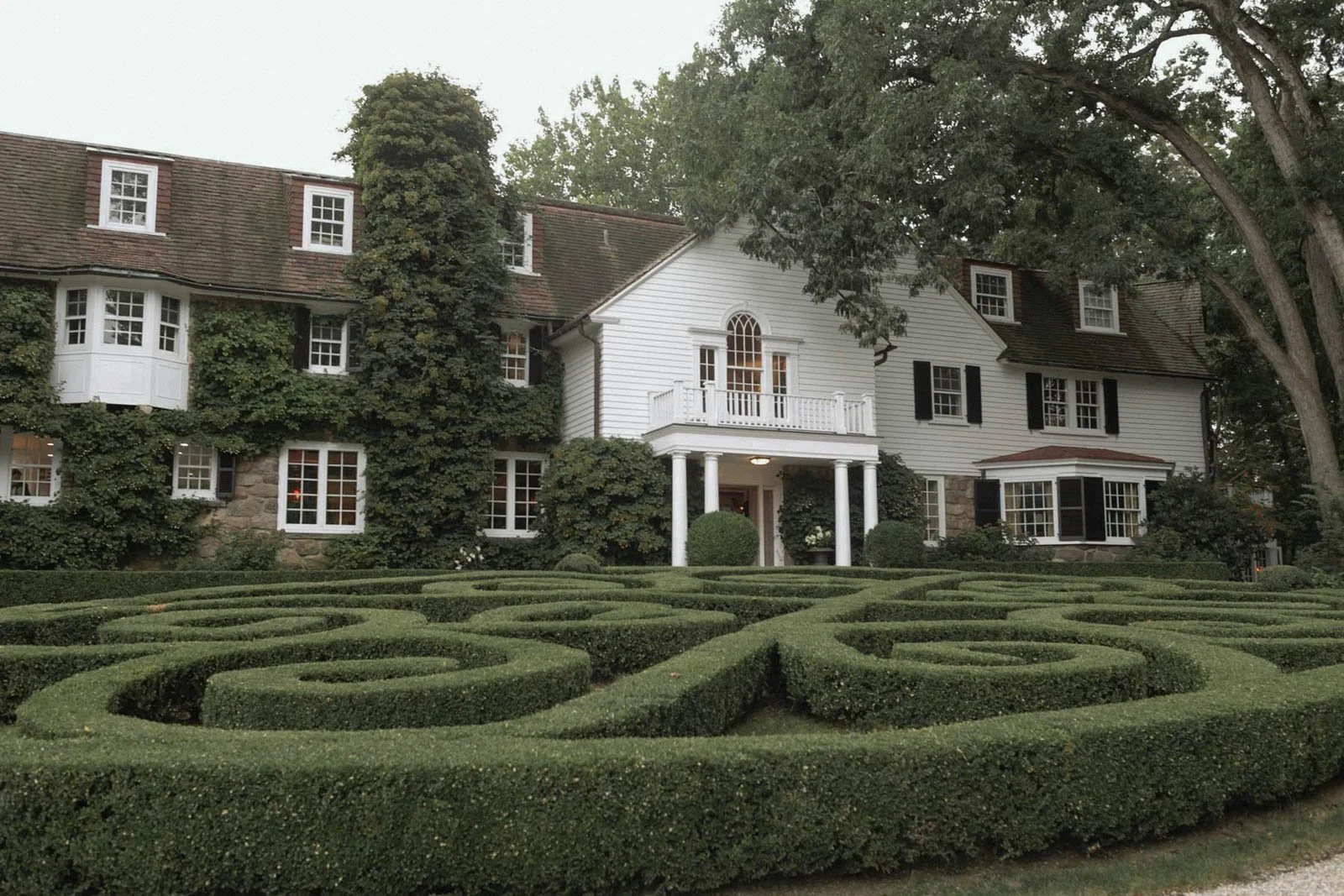 A large white house with multiple windows and black shutters, surrounded by a manicured garden with intricate hedge designs and lush trees.