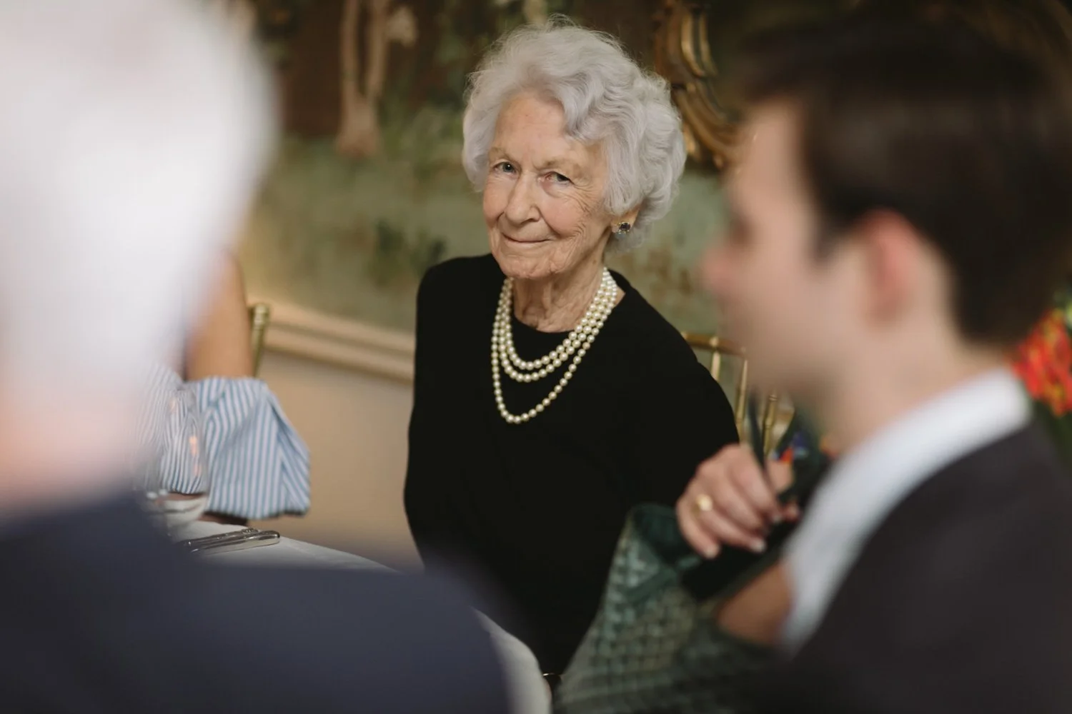 An elderly woman with white hair, wearing a black top and pearl necklace, looking at the camera during a social gathering.