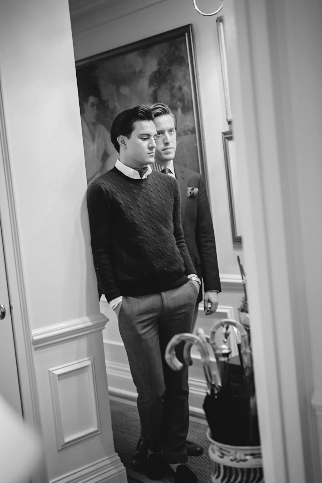 Two young men in formal attire standing in front of a mirror, one with hands in pockets, in a room with classic decor, black and white photograph.