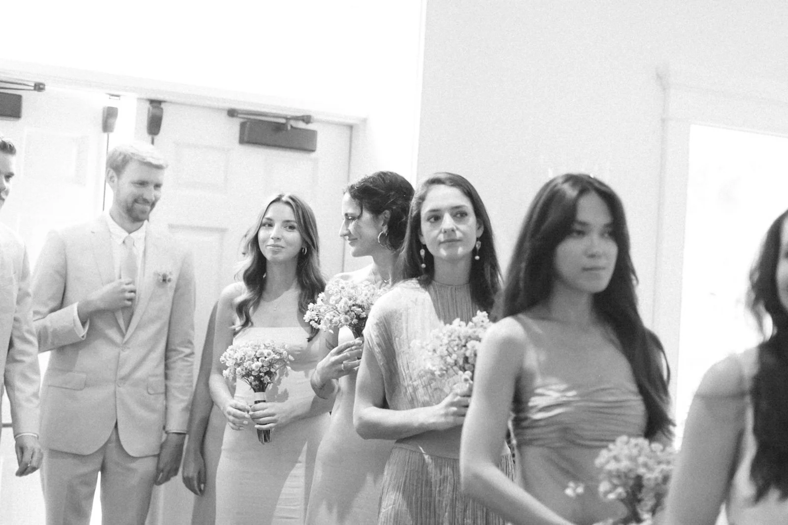 A black and white photo of a wedding processional with bridesmaids and groomsmen lined up indoors, holding bouquets, dressed in formal attire, waiting to walk down the aisle.