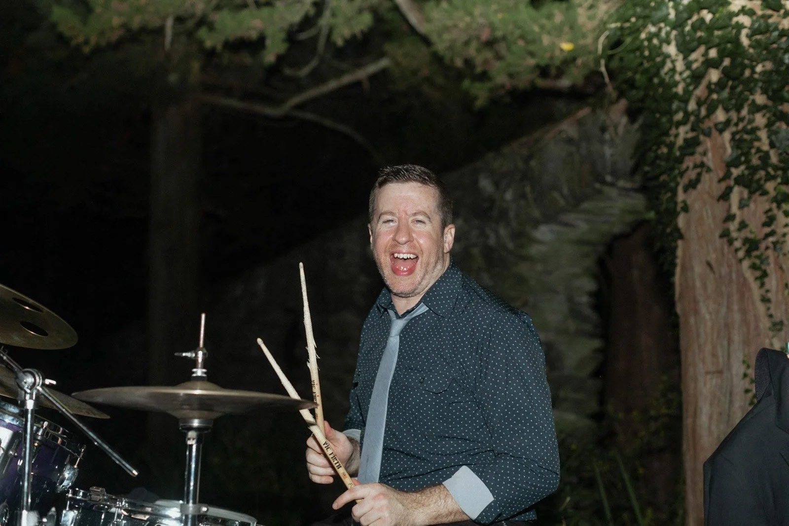 A man with short brown hair and a beard, wearing a dark blue shirt with white dots and a gray tie, holding drumsticks and smiling widely, standing near a drum set in an outdoor setting at night.