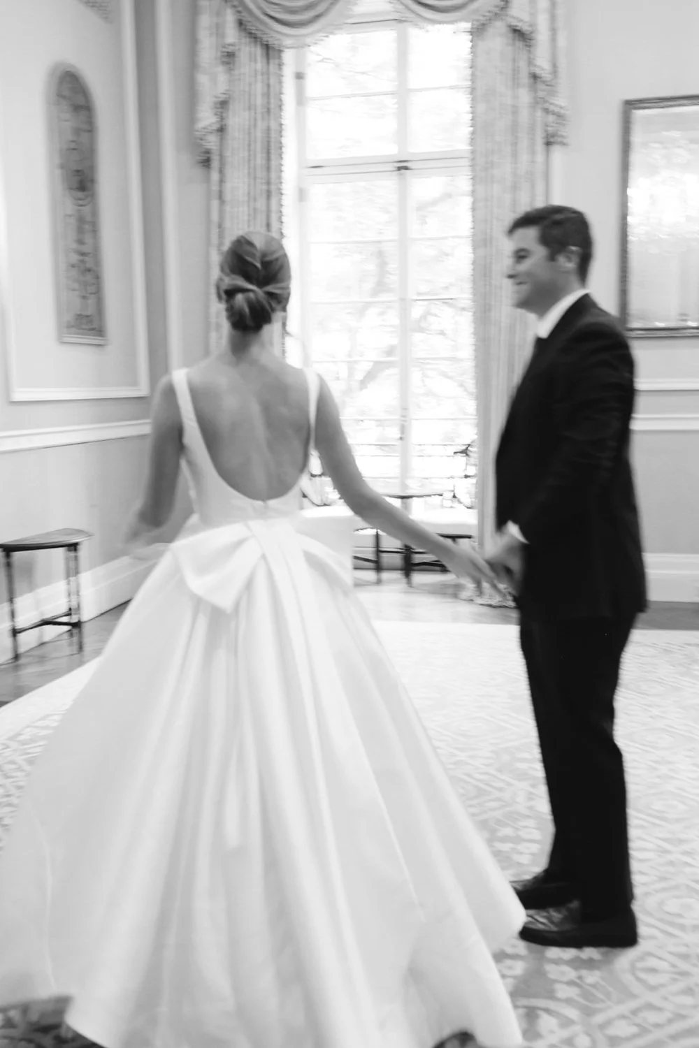 A bride in a white wedding gown is holding hands with a groom in a black tuxedo in a bright, elegantly decorated room.