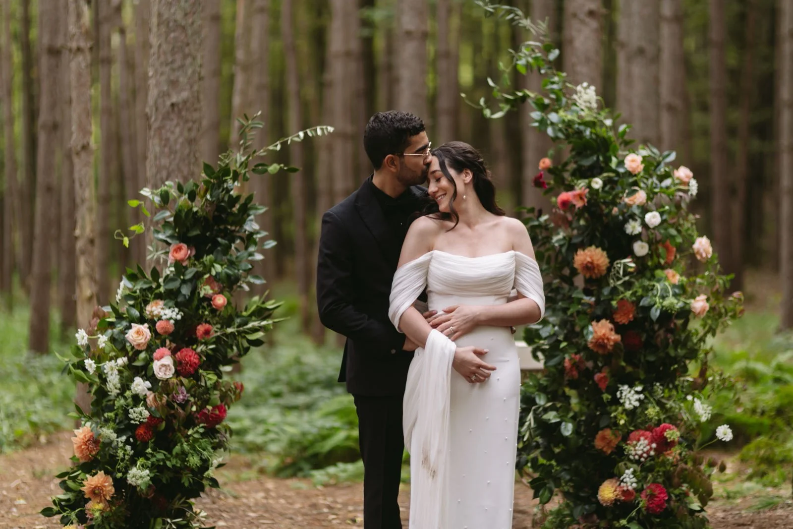 A newlywed couple in wedding attire sharing a loving moment in a forest, standing between two flower arrangements.