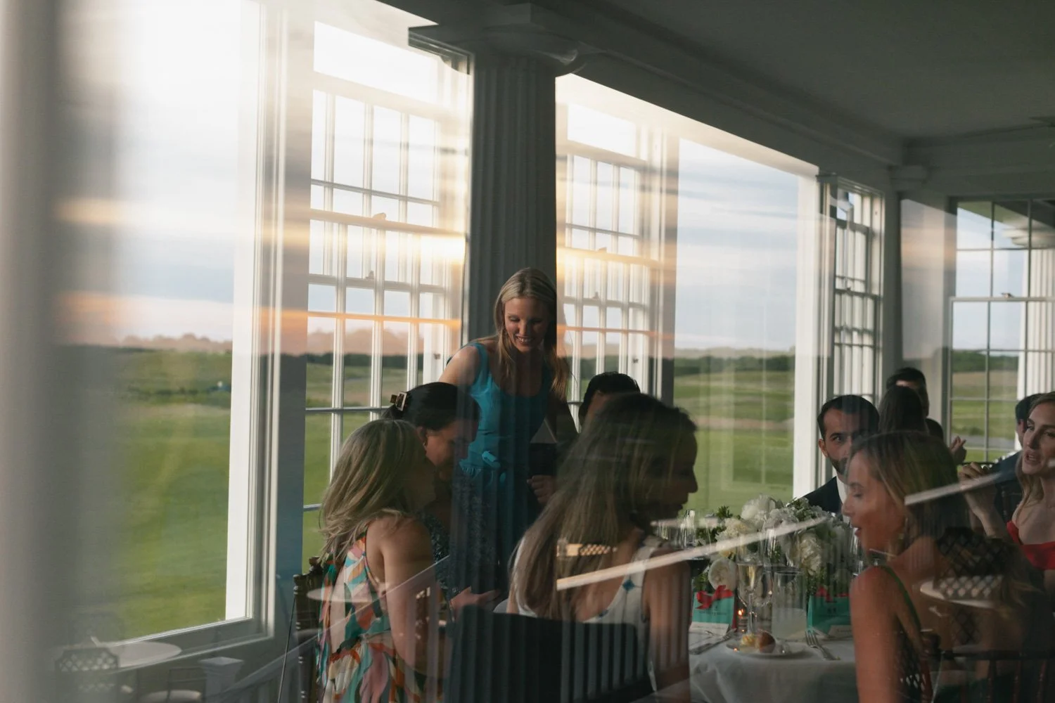 People gathered around a table inside a sunlit room with large windows overlooking a green landscape, engaging in conversation and enjoying a meal.