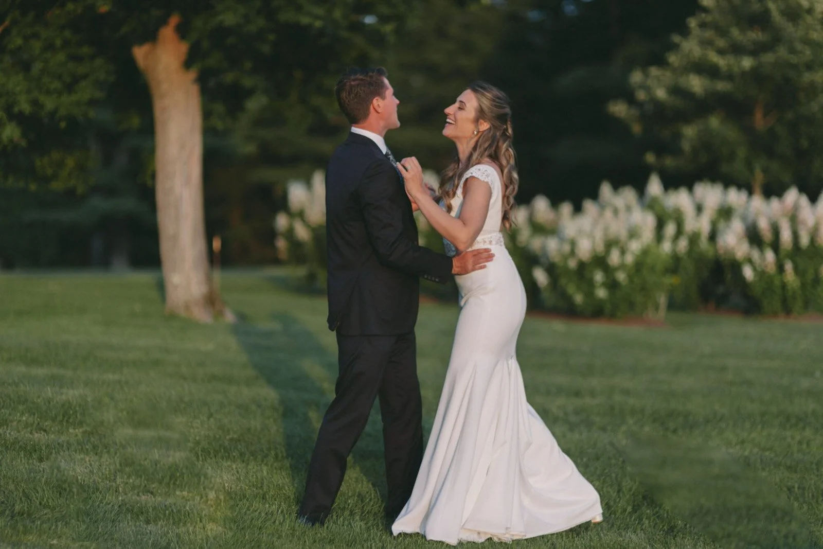 A bride and groom dancing outdoors on a wedding day, surrounded by trees and flowers, during the evening.