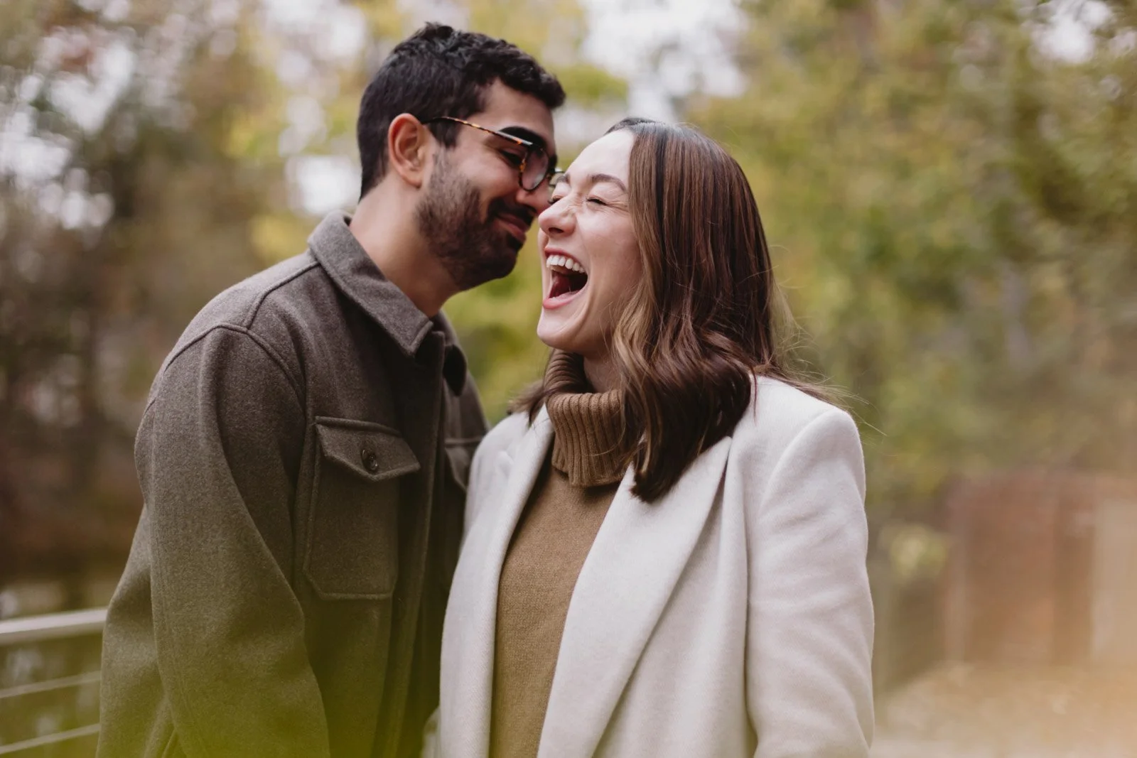 A man and woman laughing and leaning their foreheads together outdoors during fall, with autumn trees in the background.