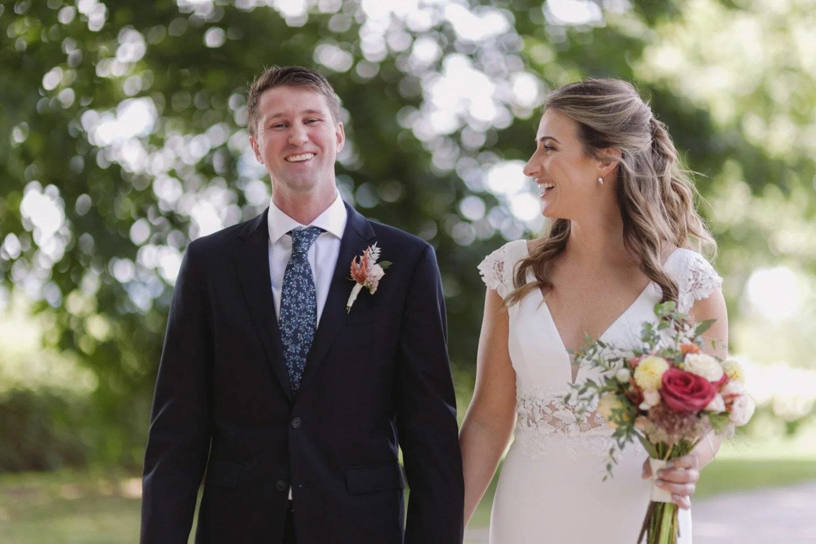 A bride and groom smiling and holding hands during their outdoor wedding ceremony, with trees and sunlight in the background.