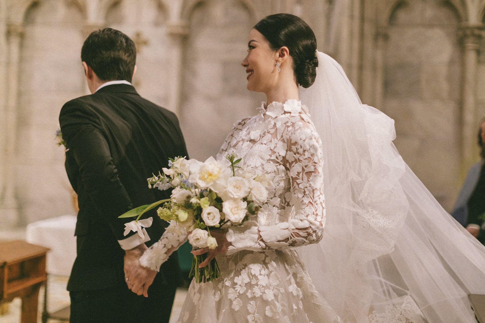 A bride wearing a lace wedding gown with a floral pattern, holding a bouquet of white flowers, standing inside a church.