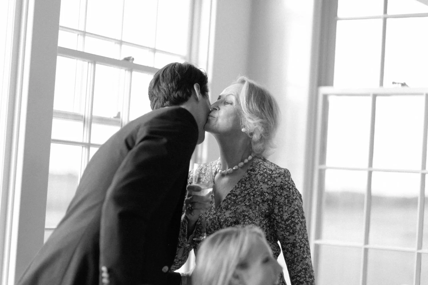 A young man in a suit kisses an older woman on the lips during a celebration in a well-lit room with large windows.
