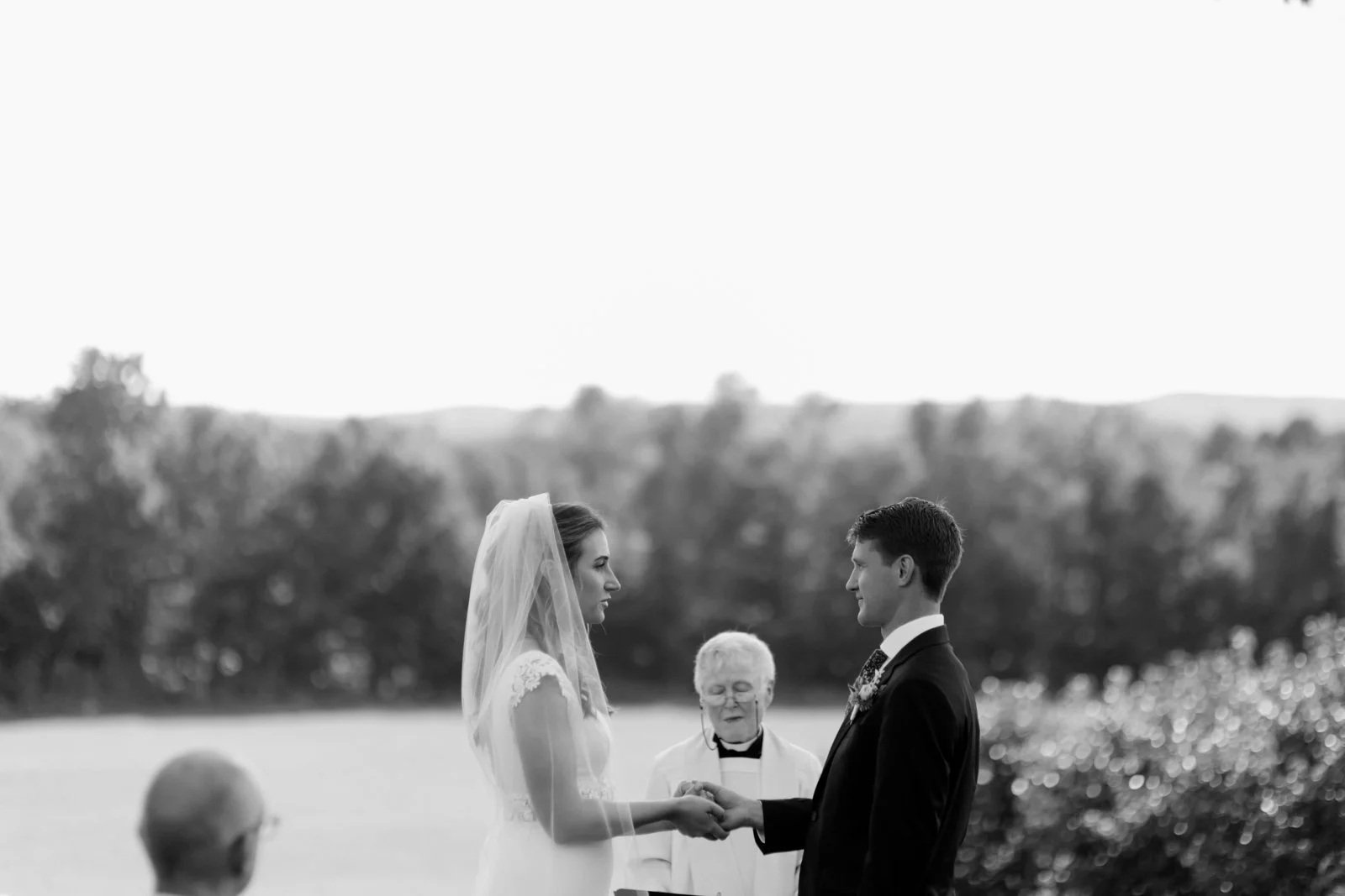 A black and white photo of a bride and groom during their outdoor wedding ceremony, holding hands and facing each other, with an officiant standing behind them. The bride wears a veil and a lace wedding dress, while the groom is in a suit and tie. Th