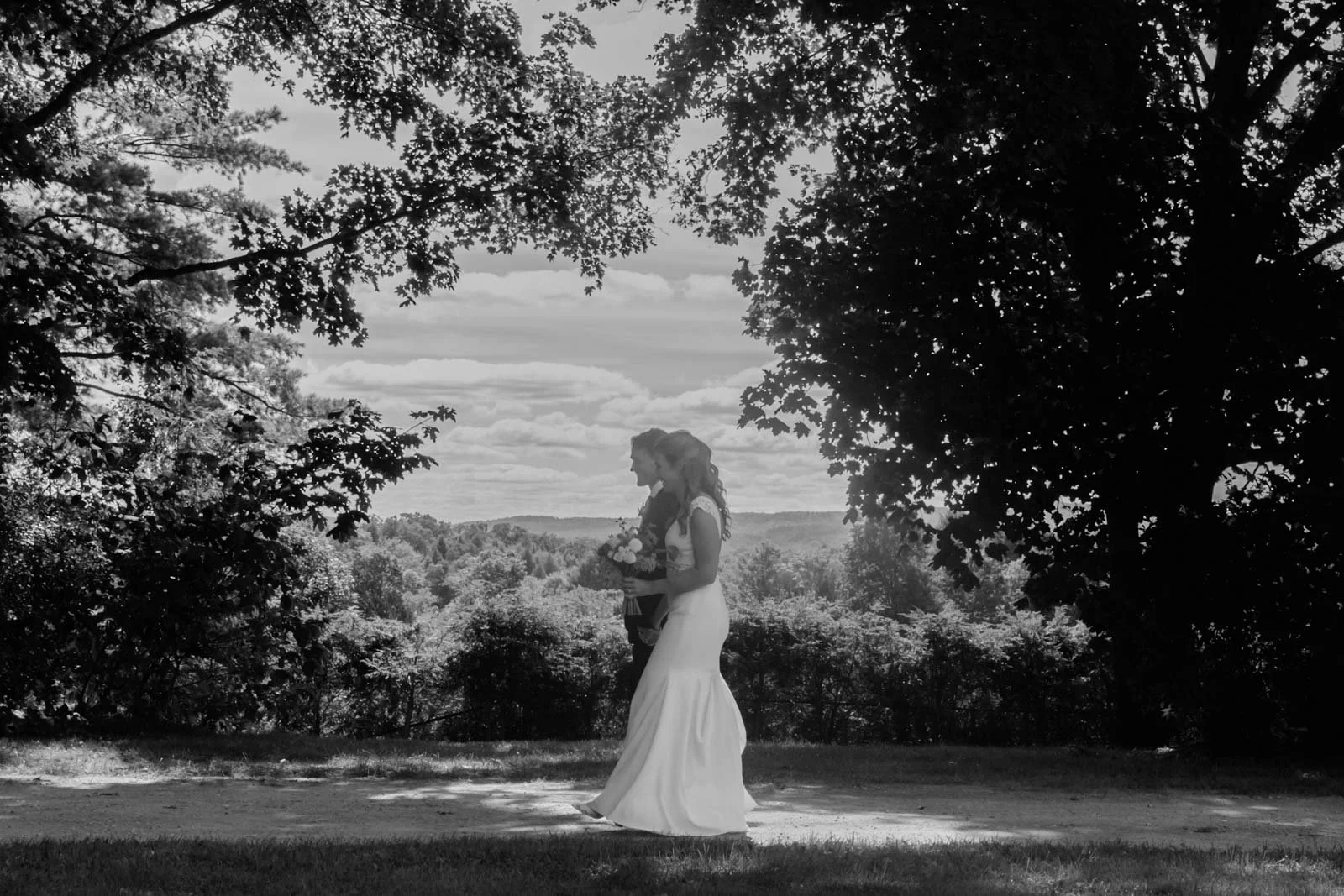 A black and white photo of a bride and groom walking outdoors beneath trees, holding a bouquet, with a scenic landscape and clouds in the background.