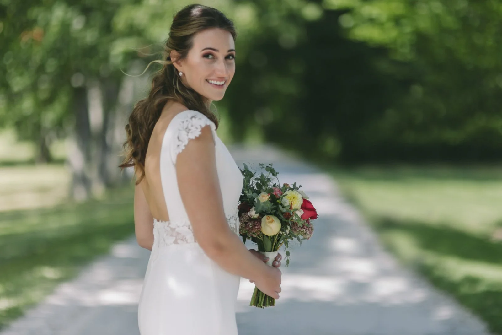 A bride standing outdoors on a sunny day, holding a bouquet of flowers, smiling and looking over her shoulder.