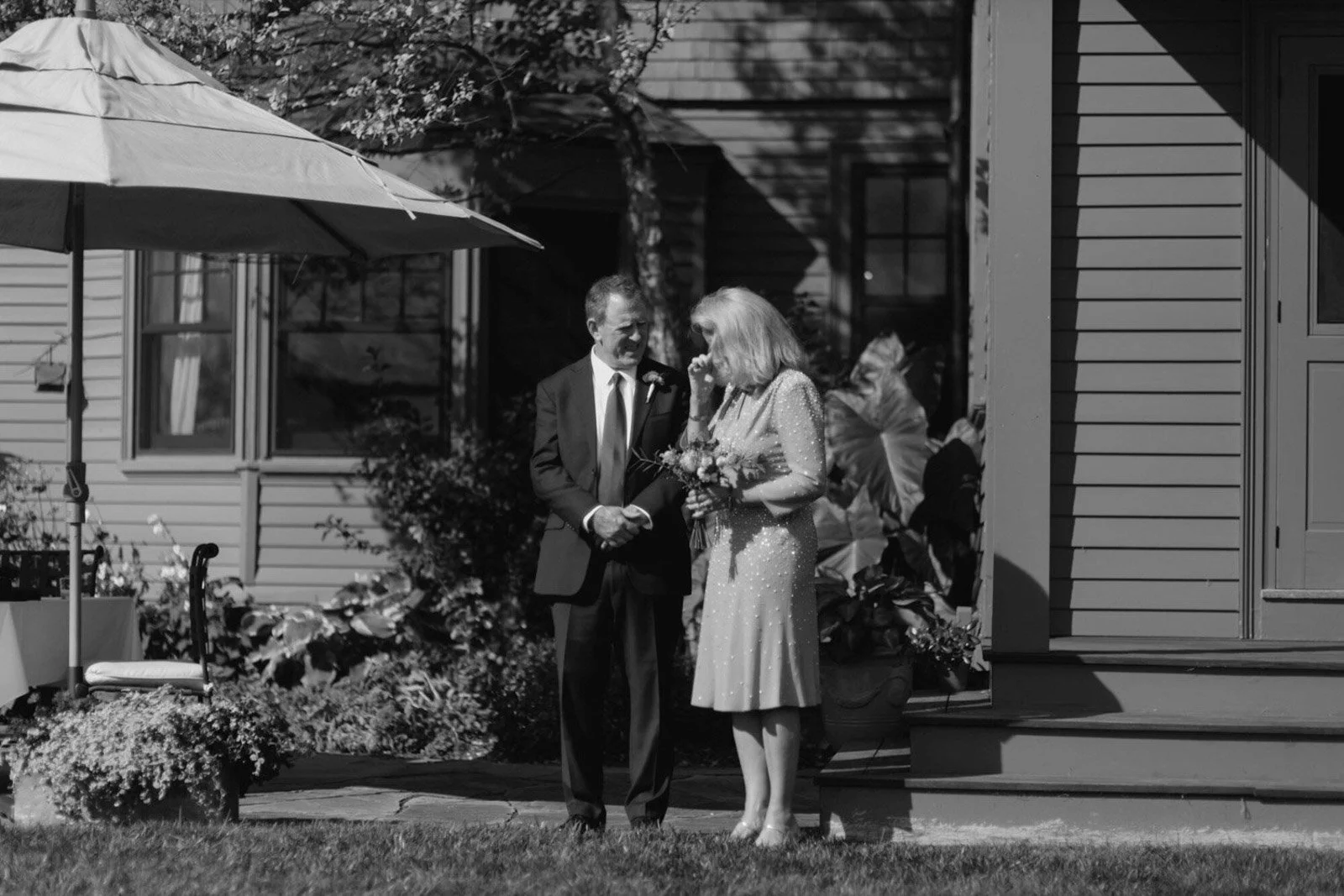 A man in a suit and a woman in a sparkling dress are standing outside a house, laughing and holding hands, during a sunny day. The woman is holding a bouquet of flowers.