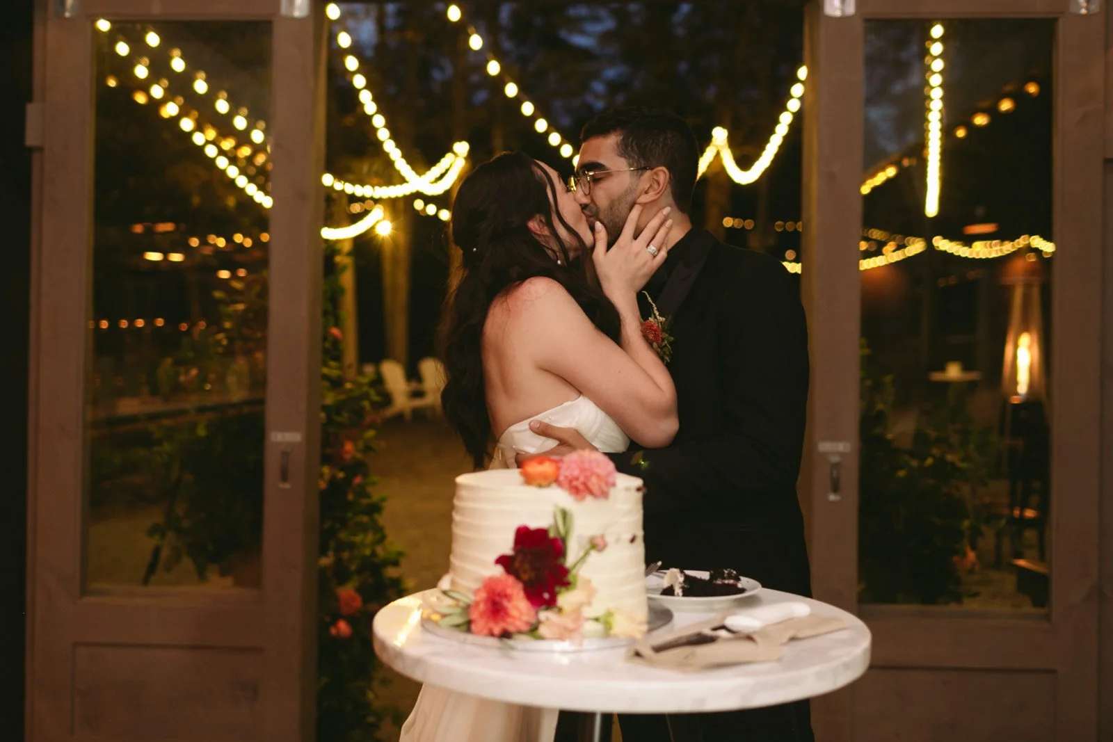 A couple sharing a kiss during their wedding reception under string lights, with a wedding cake decorated with flowers on a table in front of them.