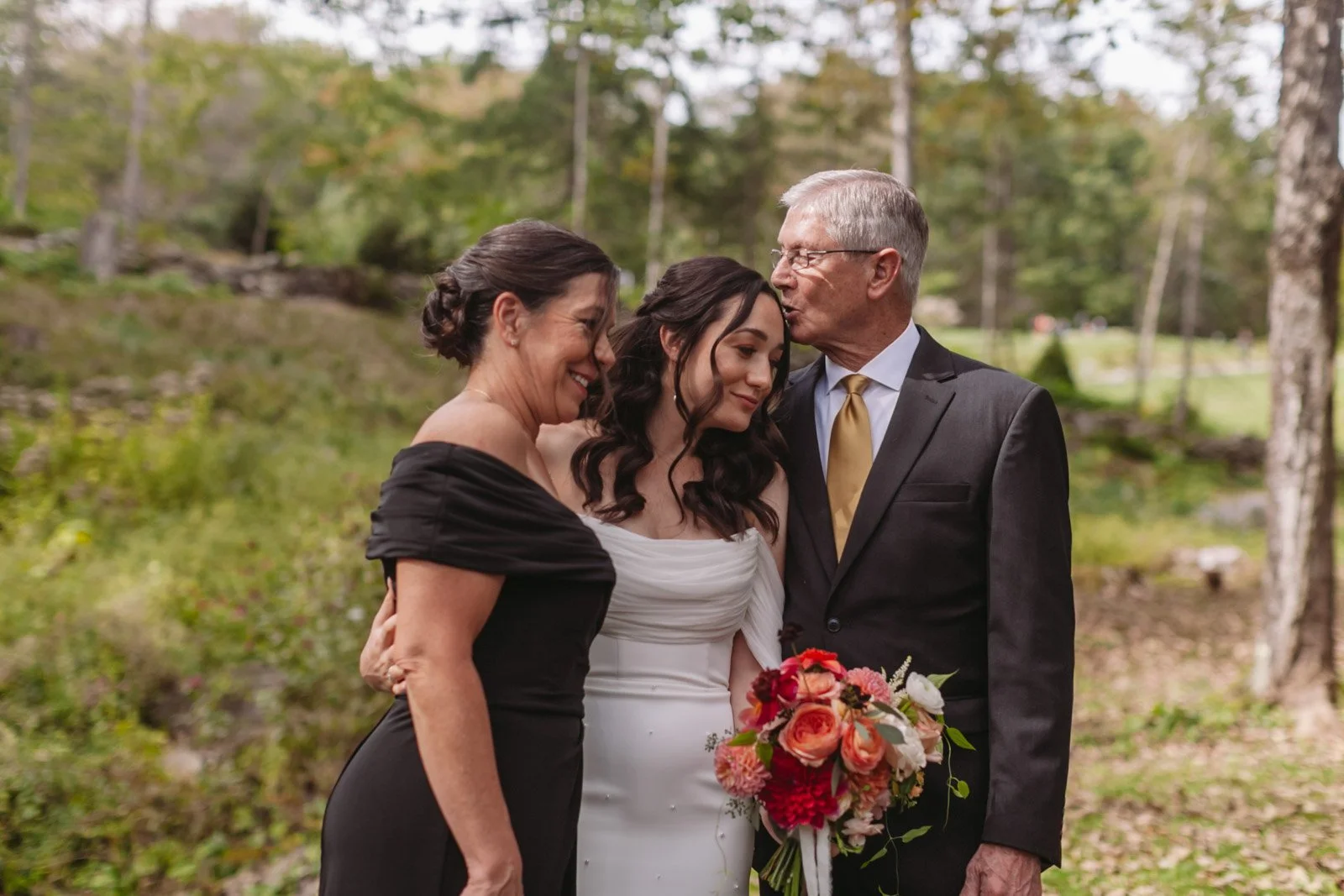 A bride with dark curly hair in a white dress holding a bouquet of pink and orange flowers, standing between an older woman in a black dress and an older man in a black suit, kissing her on the forehead in a wooded outdoor setting.