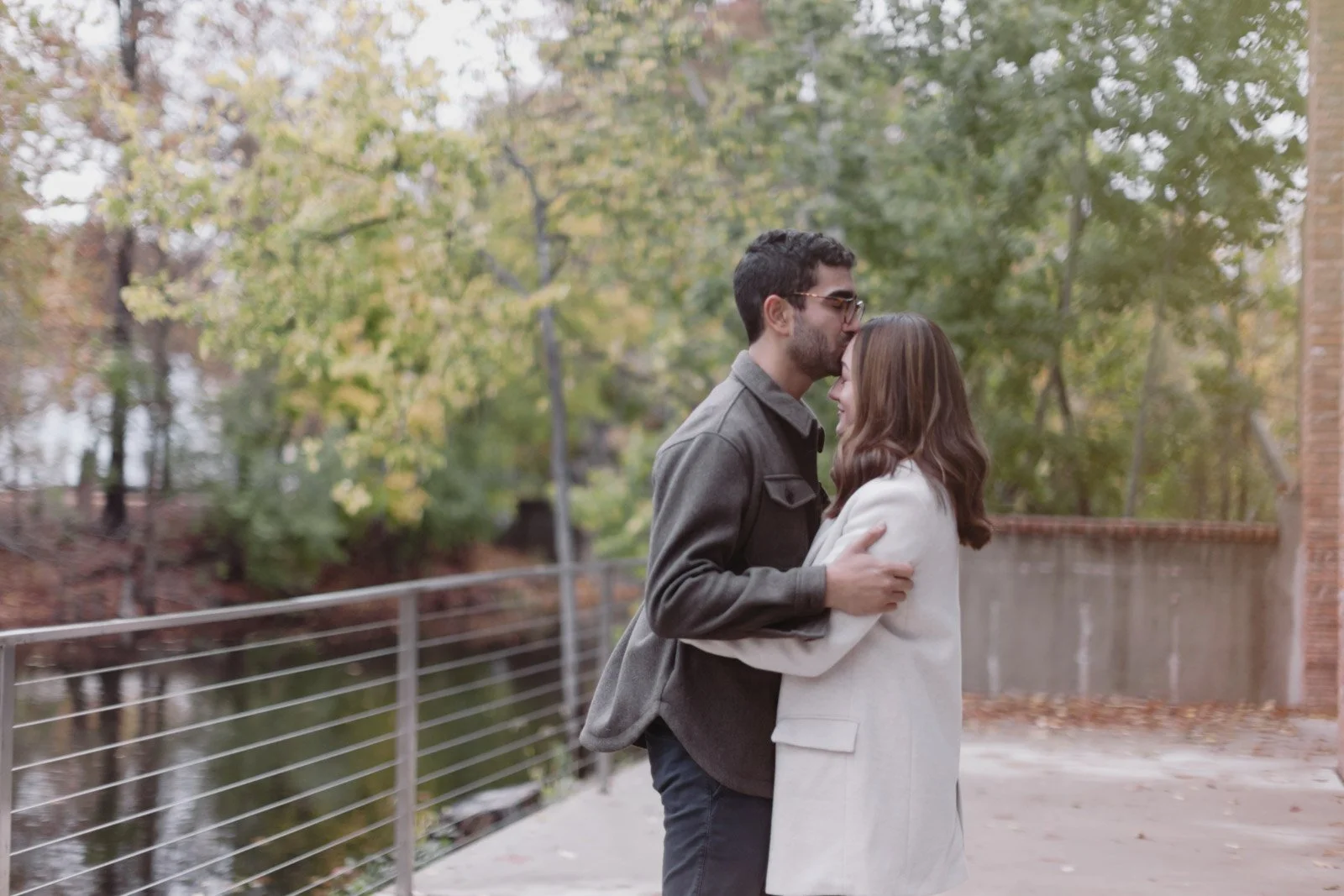 A man and woman standing close together outdoors, with the man kissing the woman's forehead. They are surrounded by fall-colored trees and a fence.