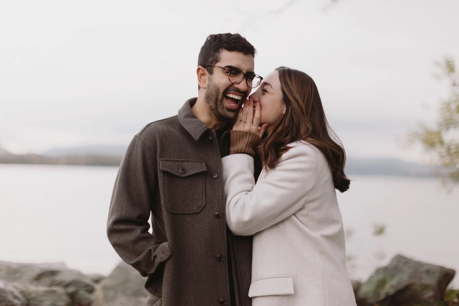 A man and woman sharing a secret by a lake, with rocks and trees in the background.