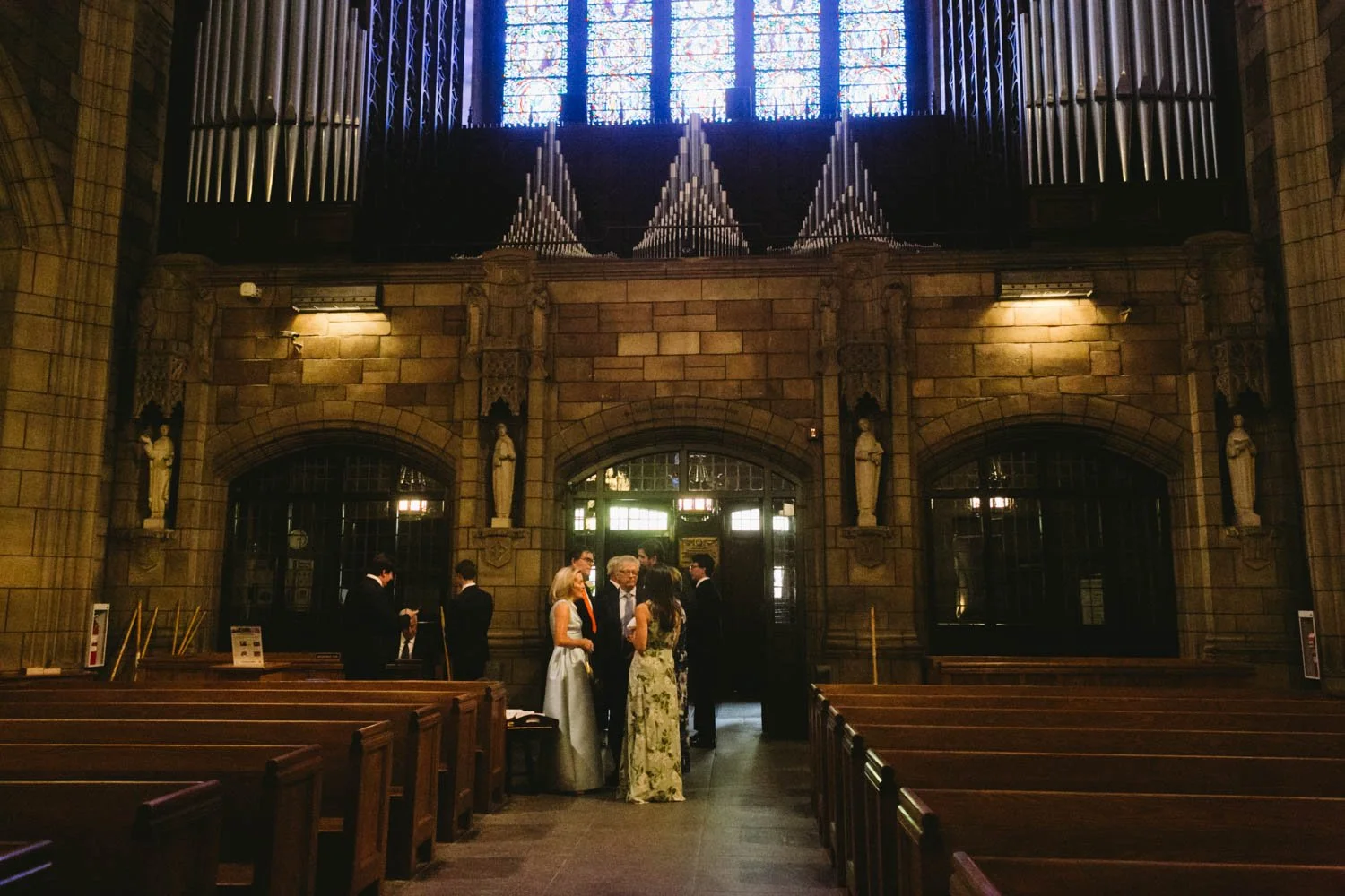 People gathering inside a church with stone walls, wooden pews, statues, stained glass windows, and large pipe organ pipes at the front.