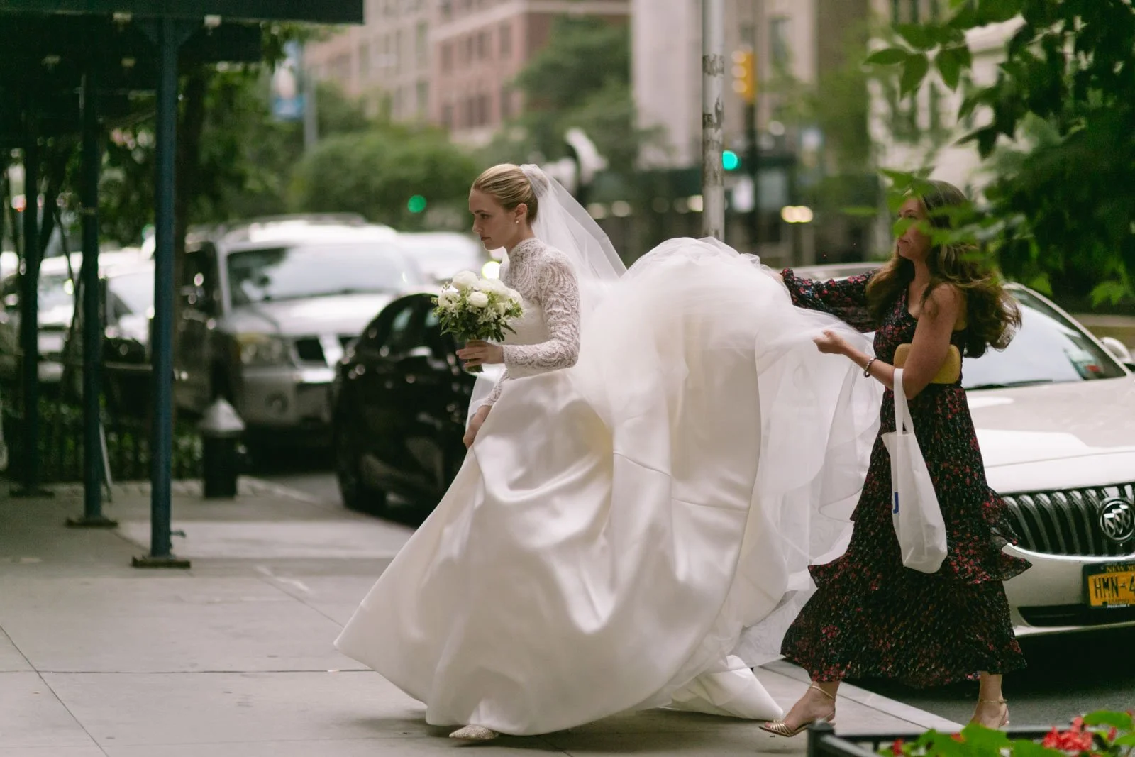 A bride in a white wedding dress holding a bouquet walking on a city sidewalk with a woman pulling her dress from behind. The scene is on a city street with cars parked along the curb and tall buildings in the background.