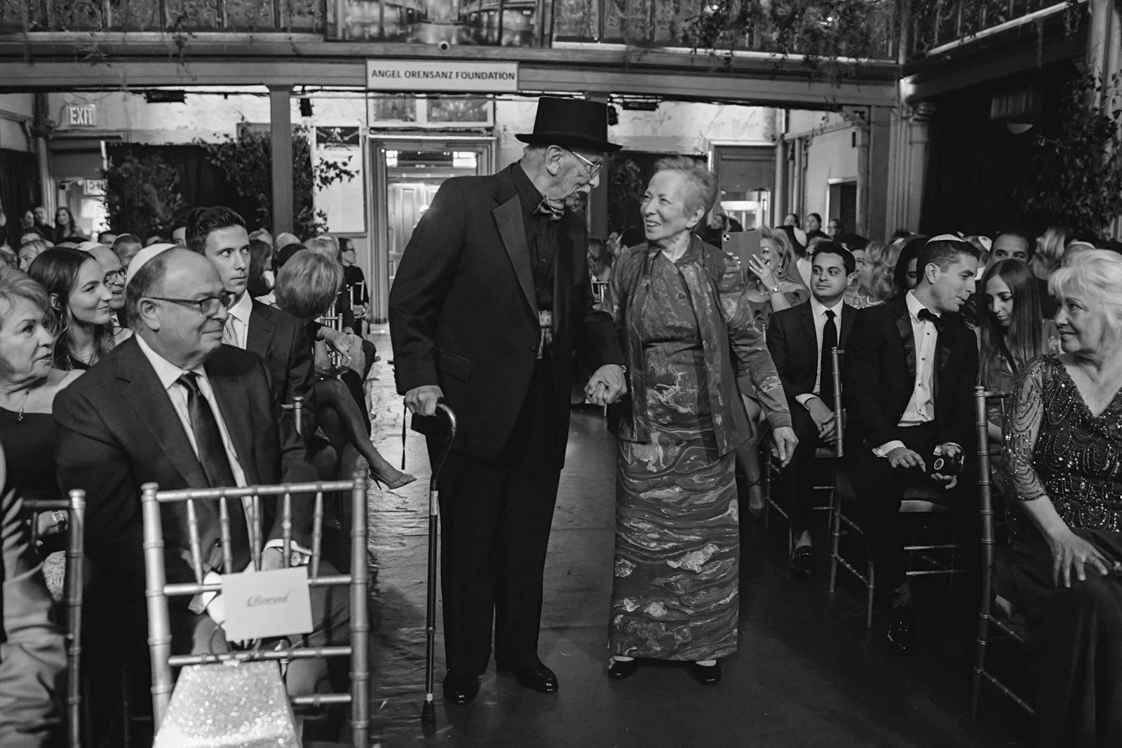 An elderly man with a cane and a top hat holding hands with an elderly woman in a patterned dress walking on a red carpet at a formal event. People are seated on either side of the carpet, dressed in formal attire, with some looking at the man and wo