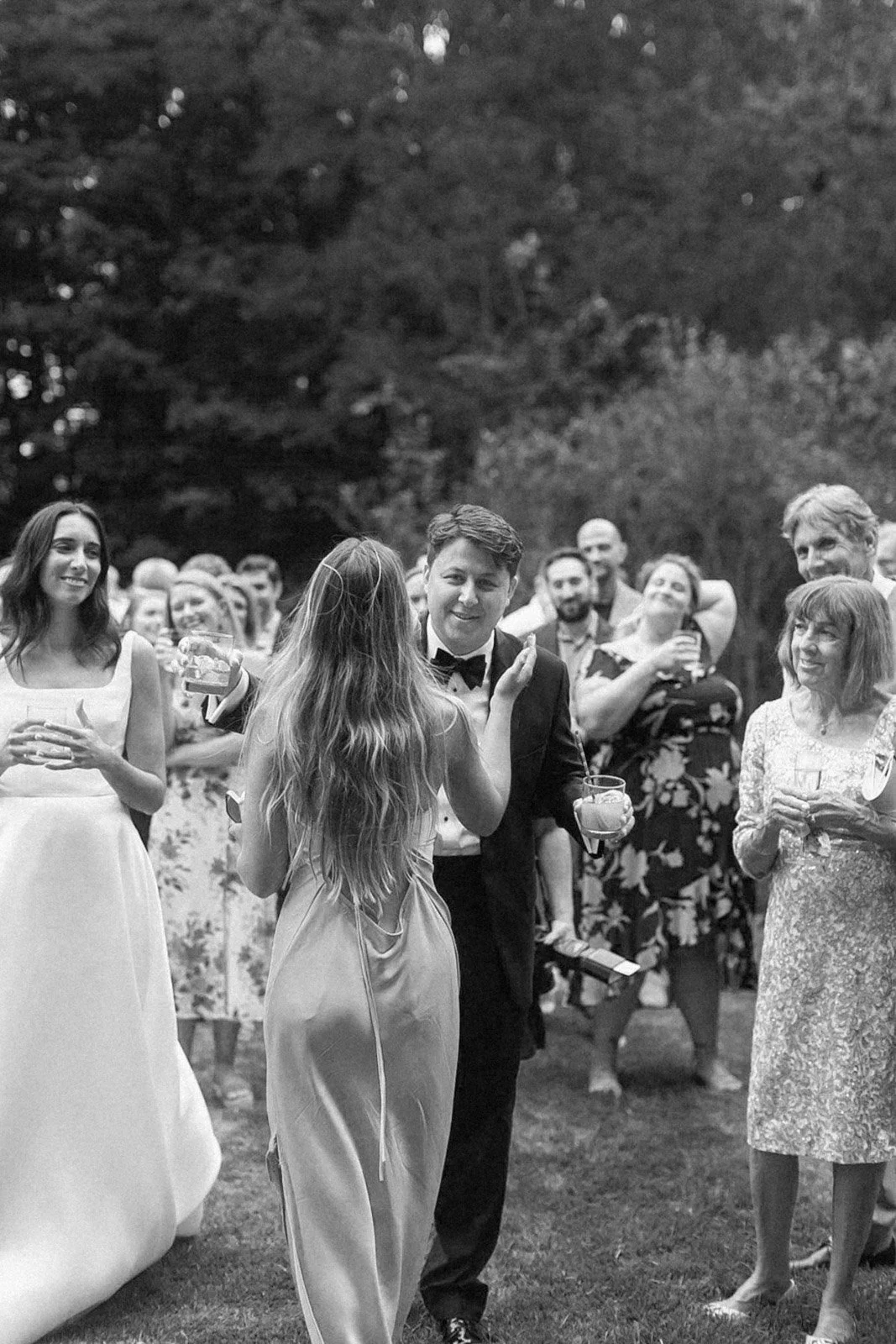 A black and white photo of a wedding reception outdoor gathering, with a woman in a white dress and a man in a tuxedo, surrounded by guests smiling and holding drinks.
