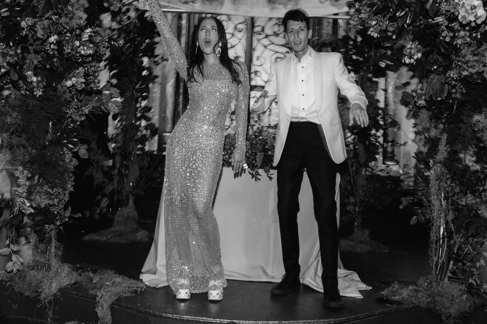 A man and woman dressed in formal attire posing on a stage or platform surrounded by floral arrangements, with the woman holding a flower and wearing a sparkly dress, and the man in a white jacket and dark pants.