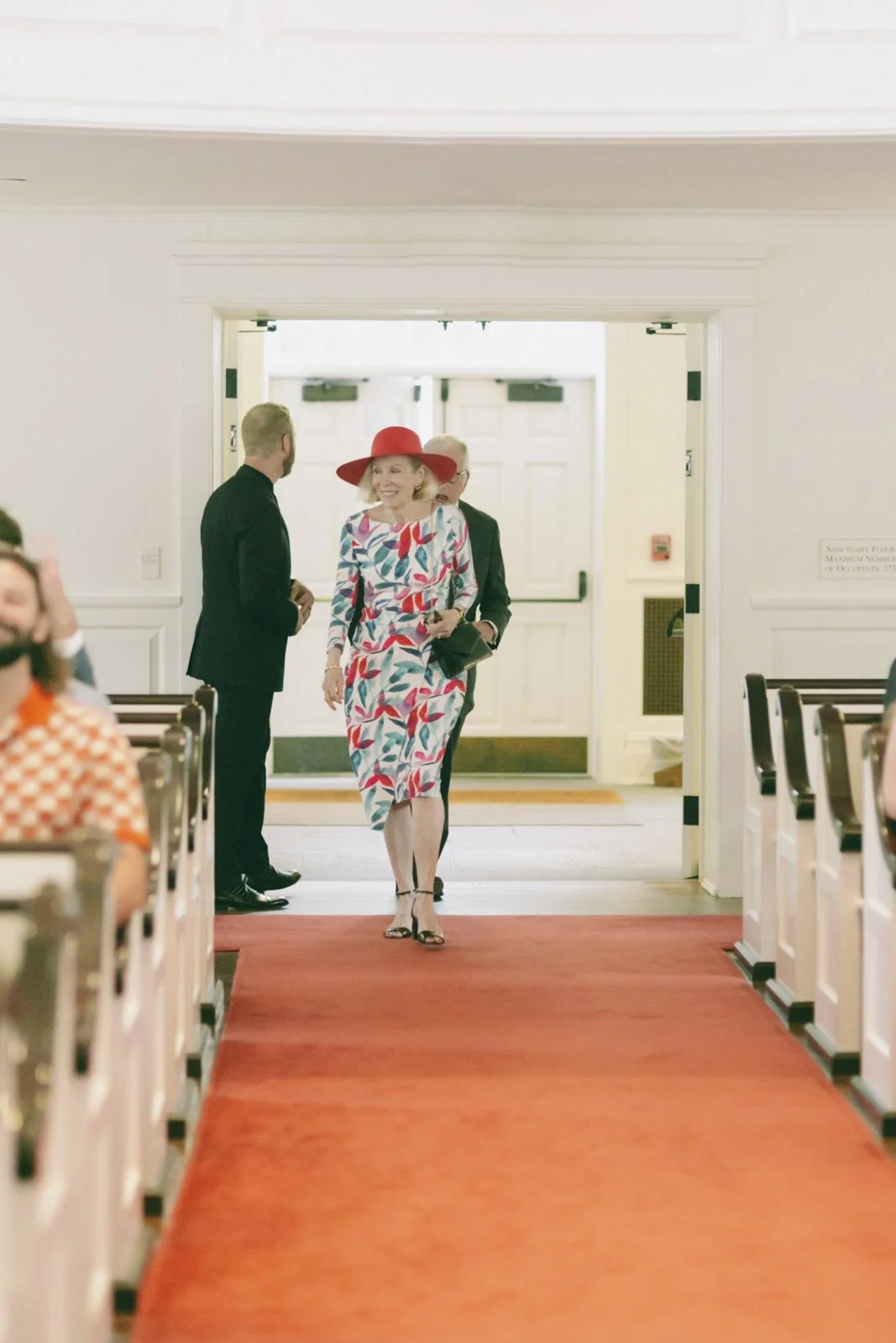 Woman in floral dress and red hat walking down the aisle of a church or hall, talking to two men in suits, while other guests are seated on either side.