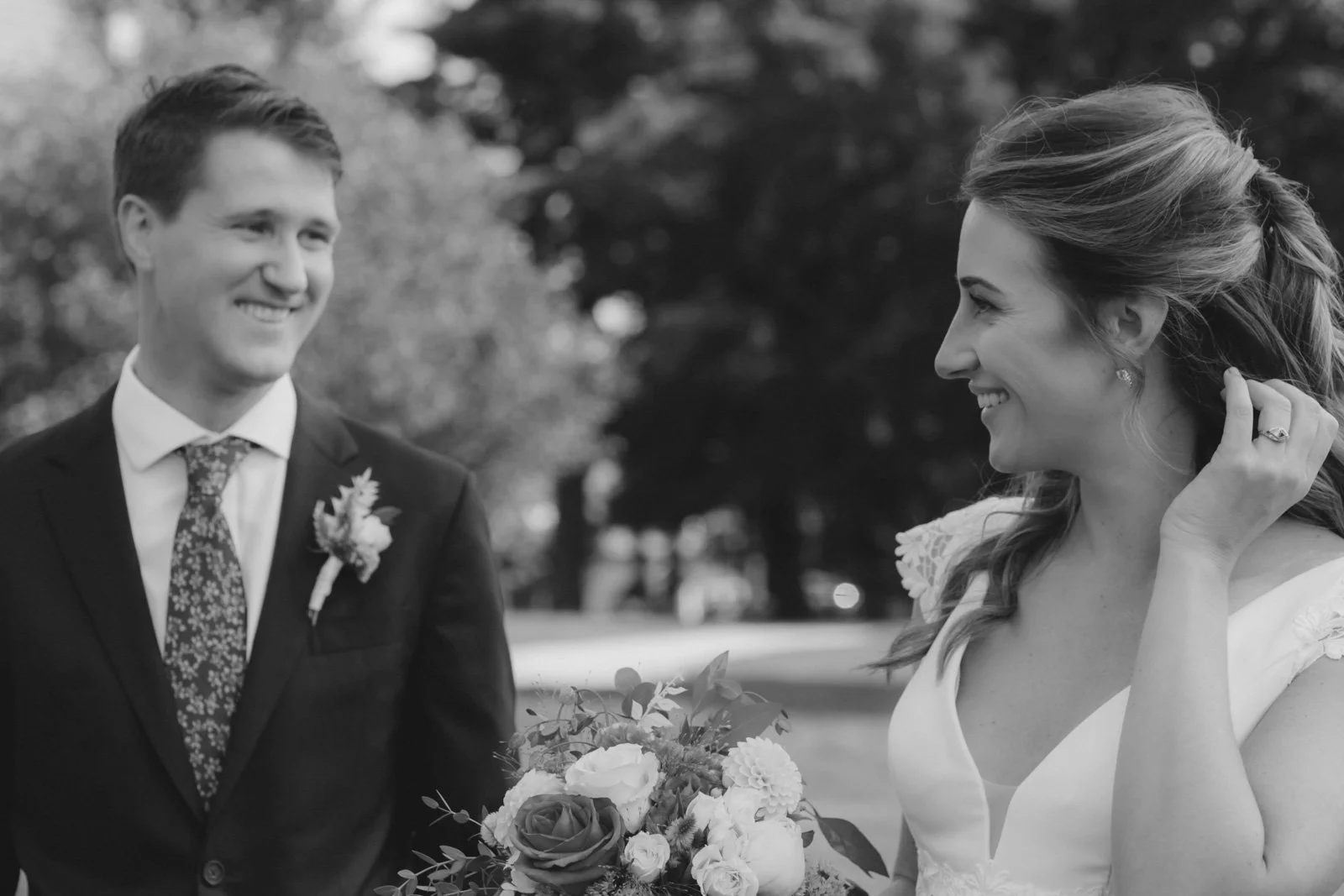 A smiling groom in a suit with a boutonnière looks at a smiling bride holding a bouquet with a garden in the background.