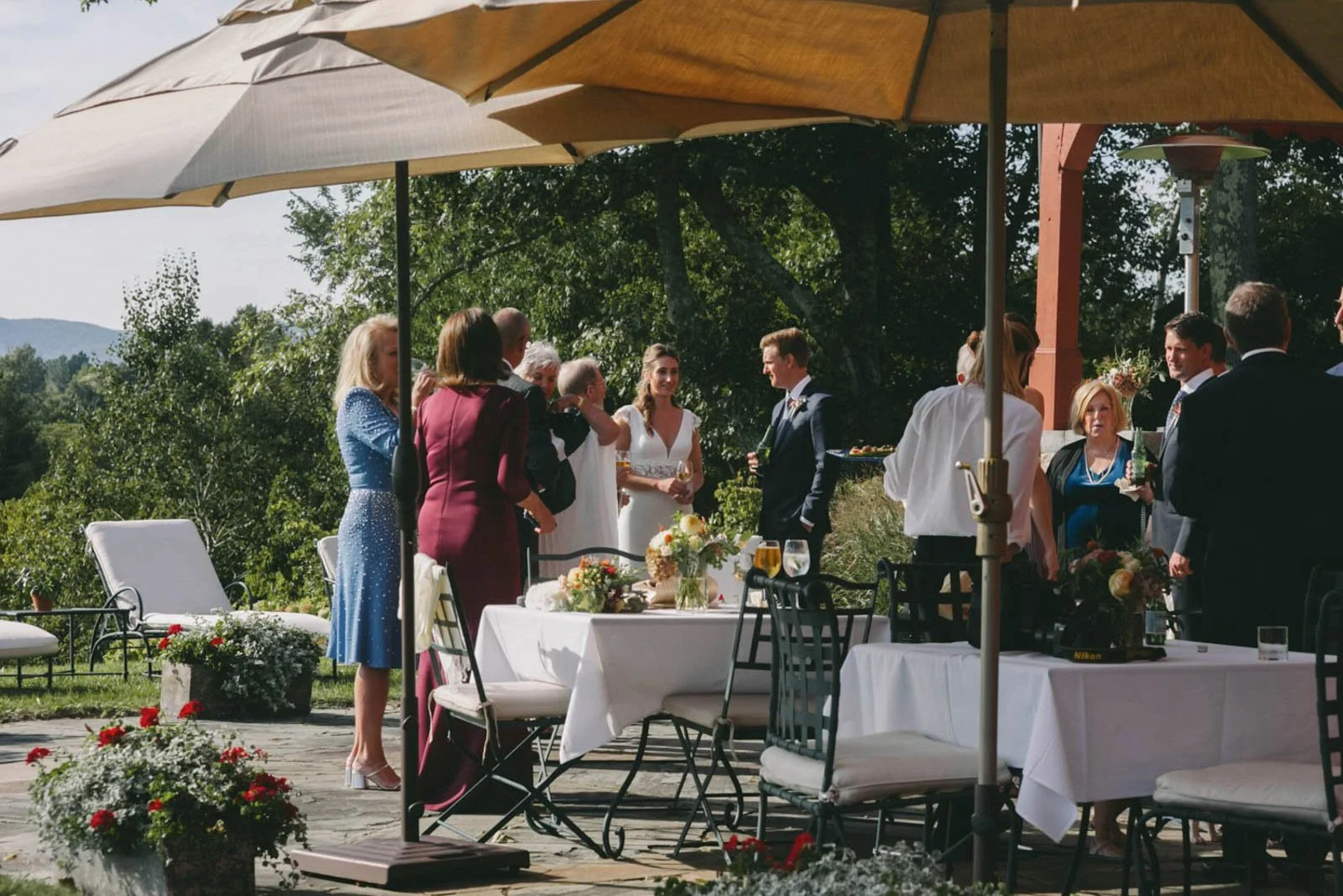Outdoor wedding reception with guests mingling under large beige umbrellas, decorated tables with flowers, and a bride and groom standing together, smiling.