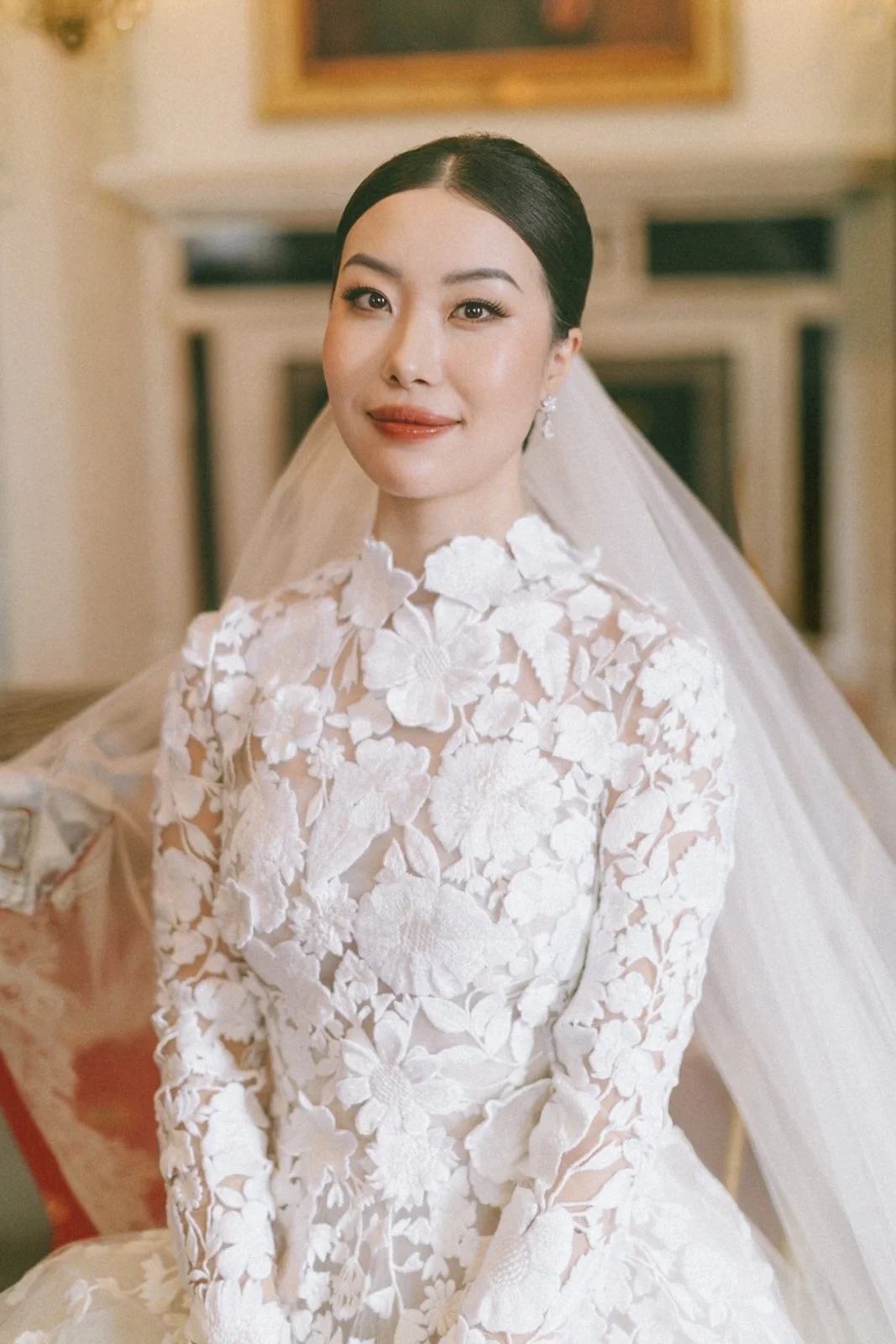 A bride with dark hair in a sleek bun, wearing a white lace wedding gown with floral embroidery and a veil, smiling in an indoor setting.