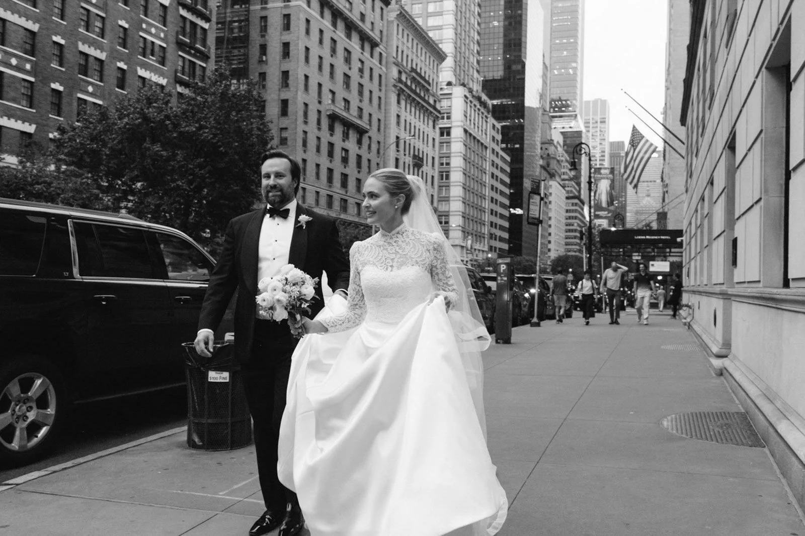 Black and white photo of a bride and groom walking on a city sidewalk, with tall buildings and a few pedestrians in the background. The bride is holding her dress and a bouquet, and the groom is in a tuxedo.