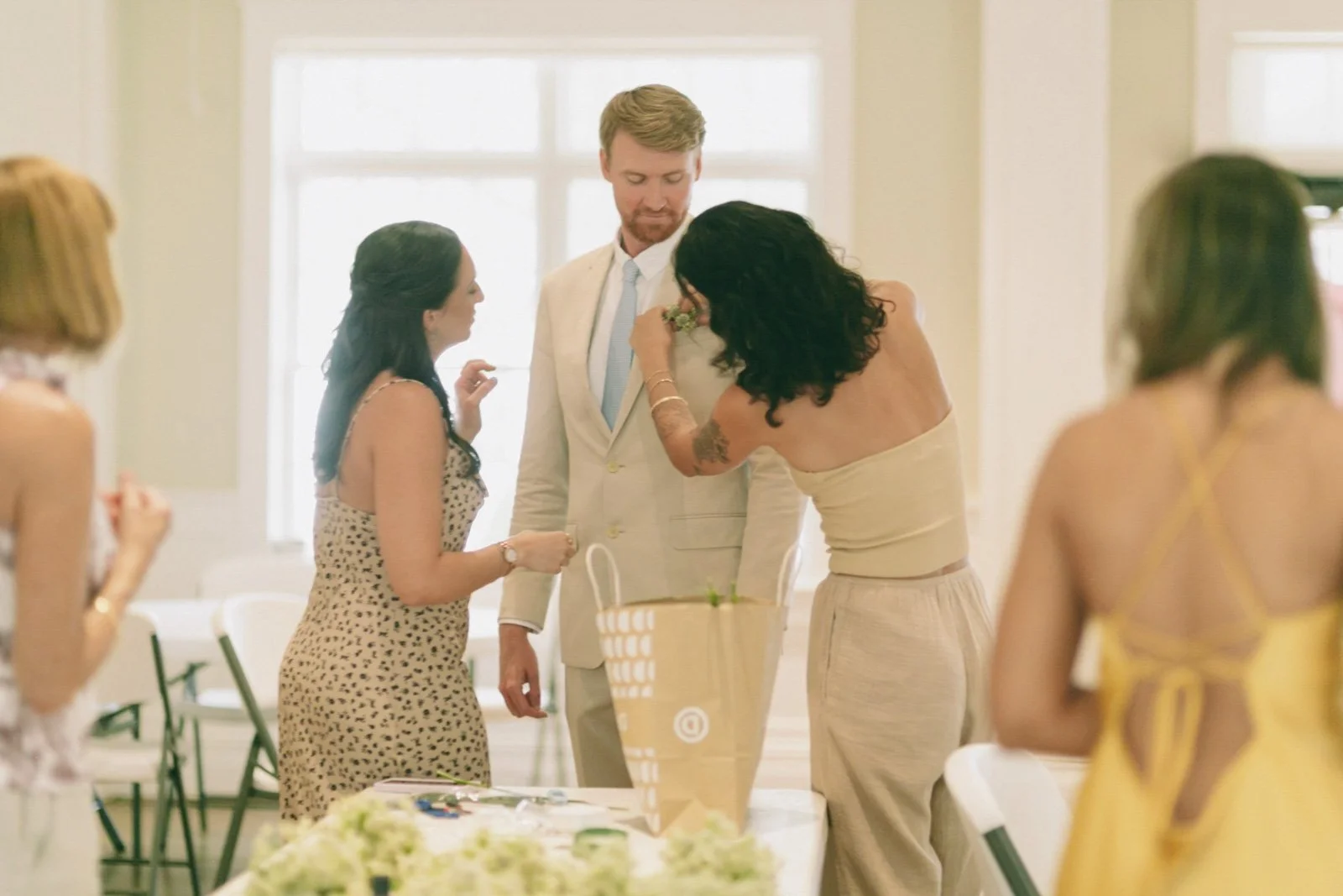 People at a celebration, with a woman pinning a boutonniere on a man's jacket at a decorated indoor event.