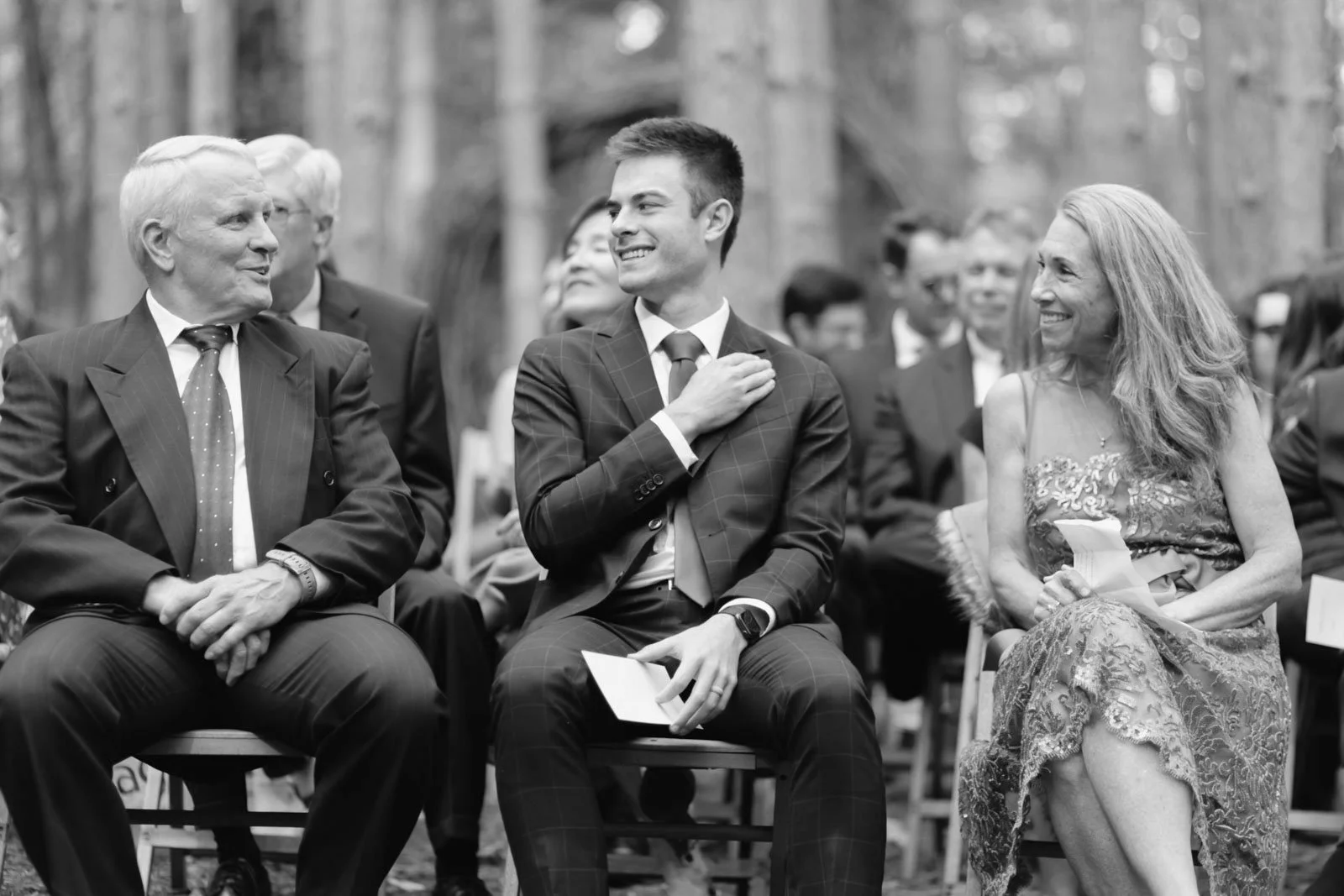 People attending an outdoor formal event, sitting on chairs, with a man in a suit and woman in an embroidered dress smiling and engaging in conversation.