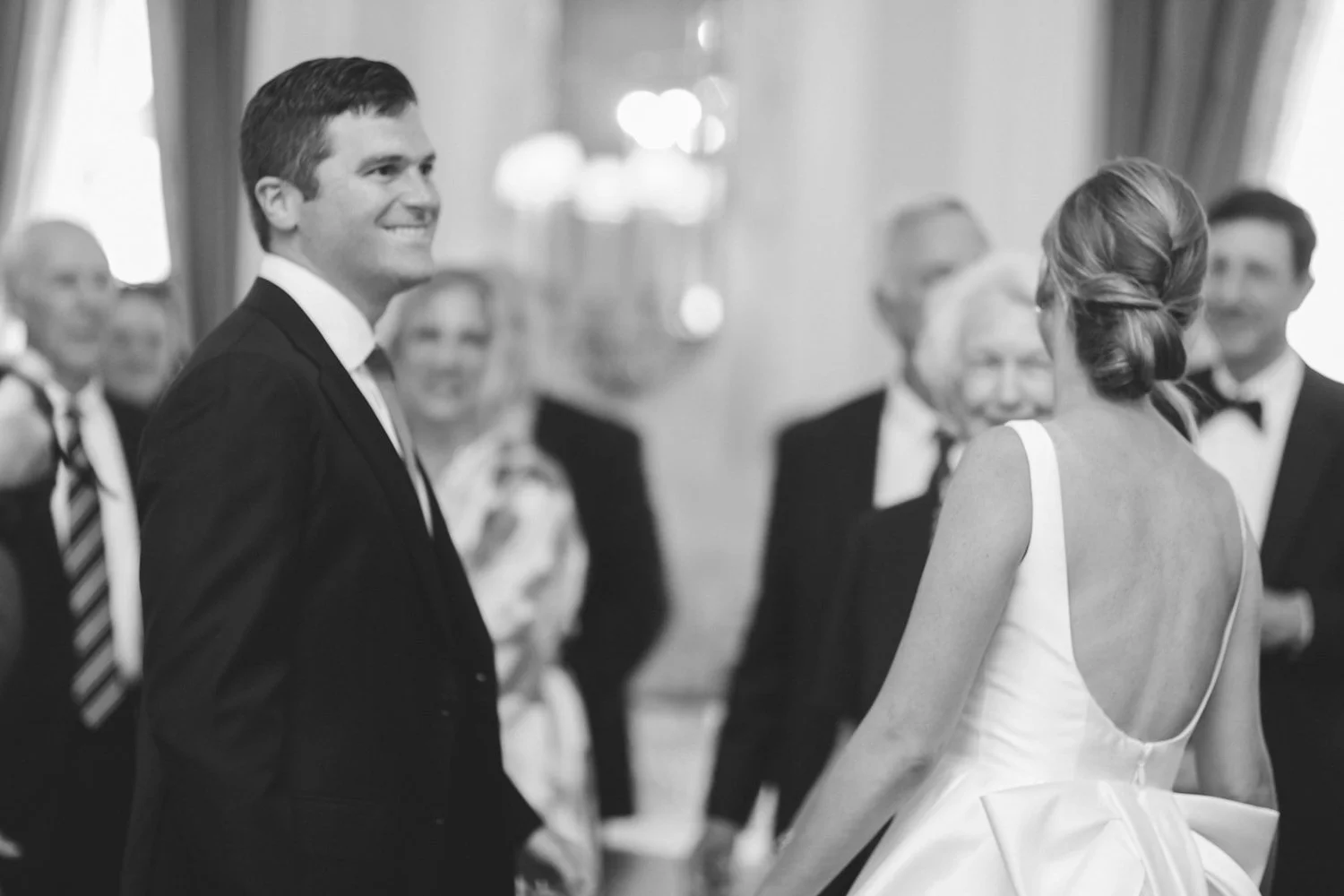 A wedding ceremony with a bride and groom holding hands, looking at each other, surrounded by older guests, in black and white.