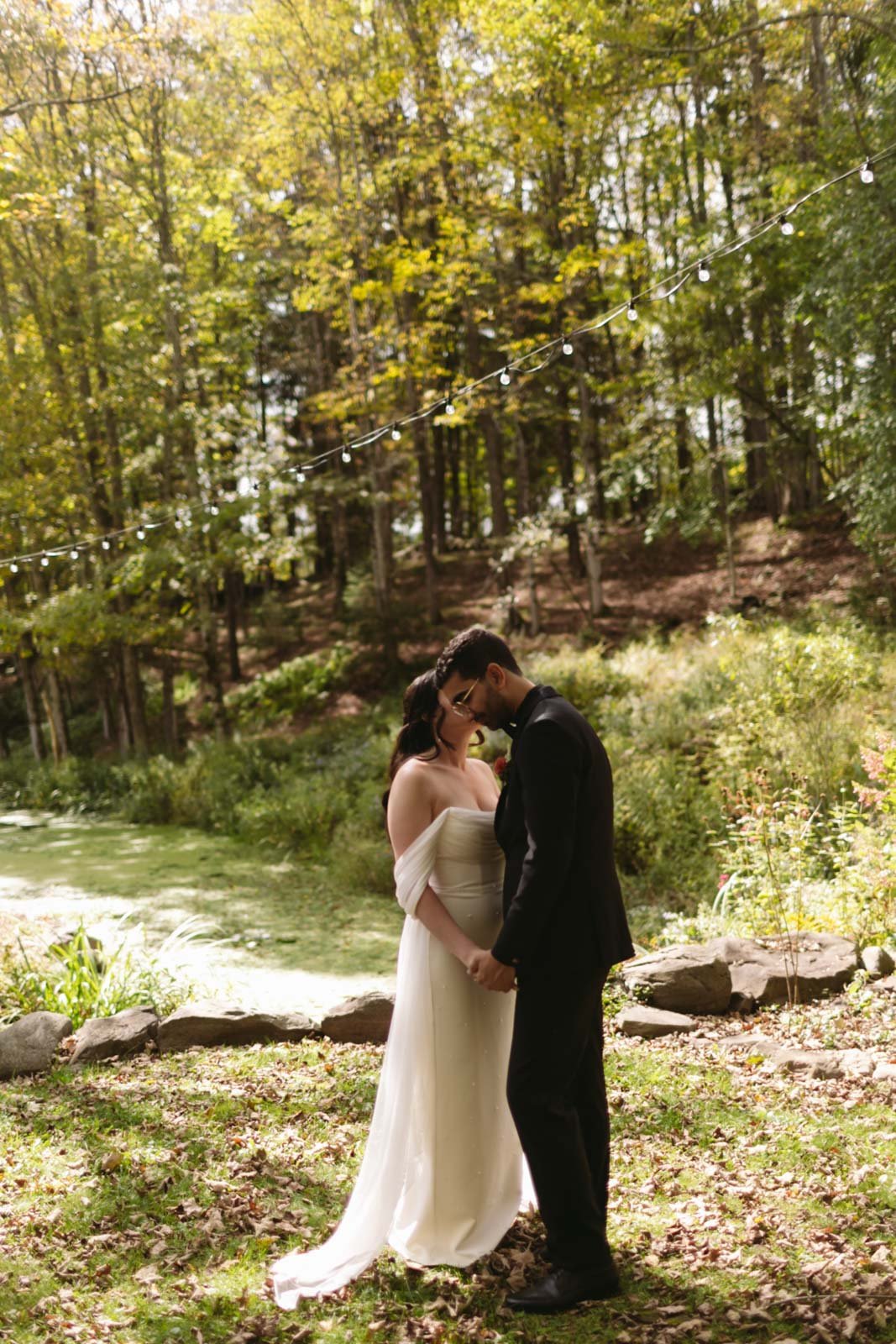 A couple dressed in wedding attire holding hands and leaning their foreheads together outdoors in a wooded area with string lights overhead.
