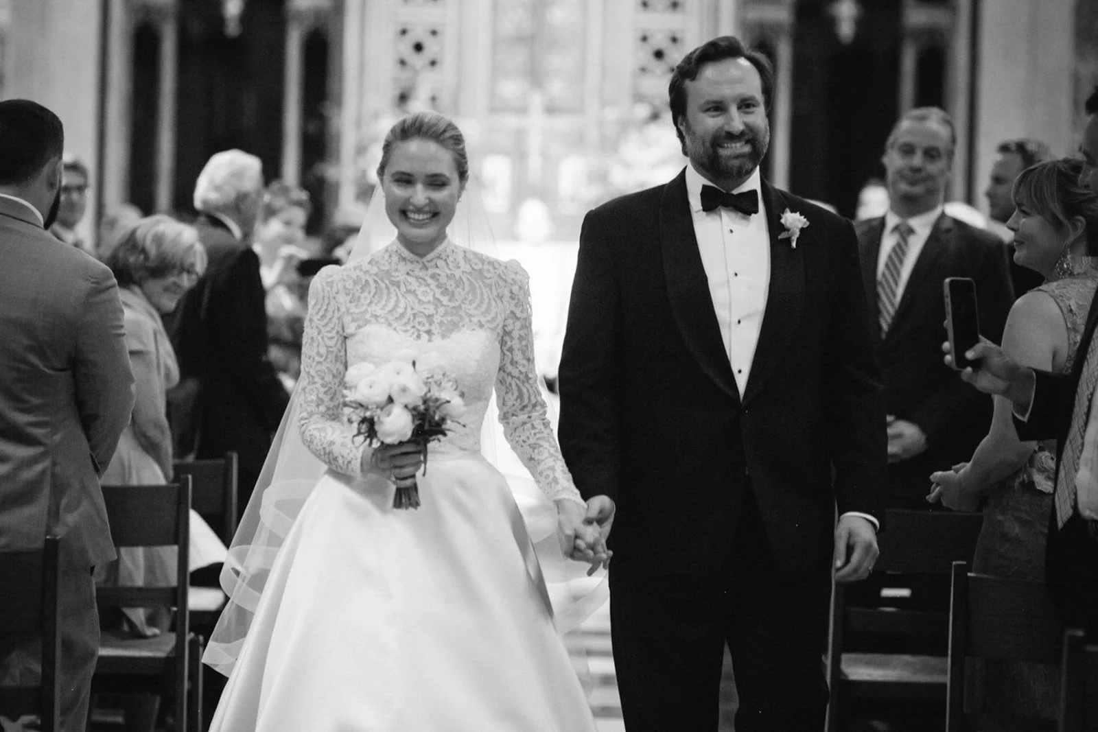 A black-and-white photo of a wedding ceremony with a bride and groom walking down the aisle, holding hands, surrounded by seated guests in a church.