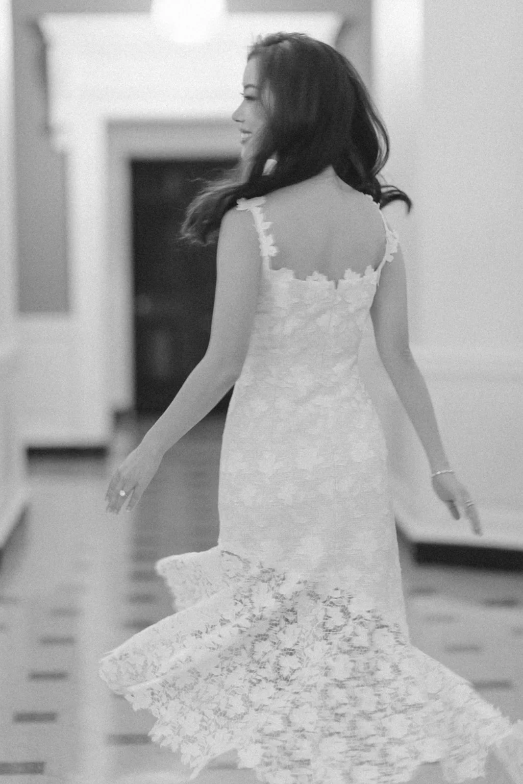 A woman in a lace dress walking through a hallway, smiling and looking to the side.