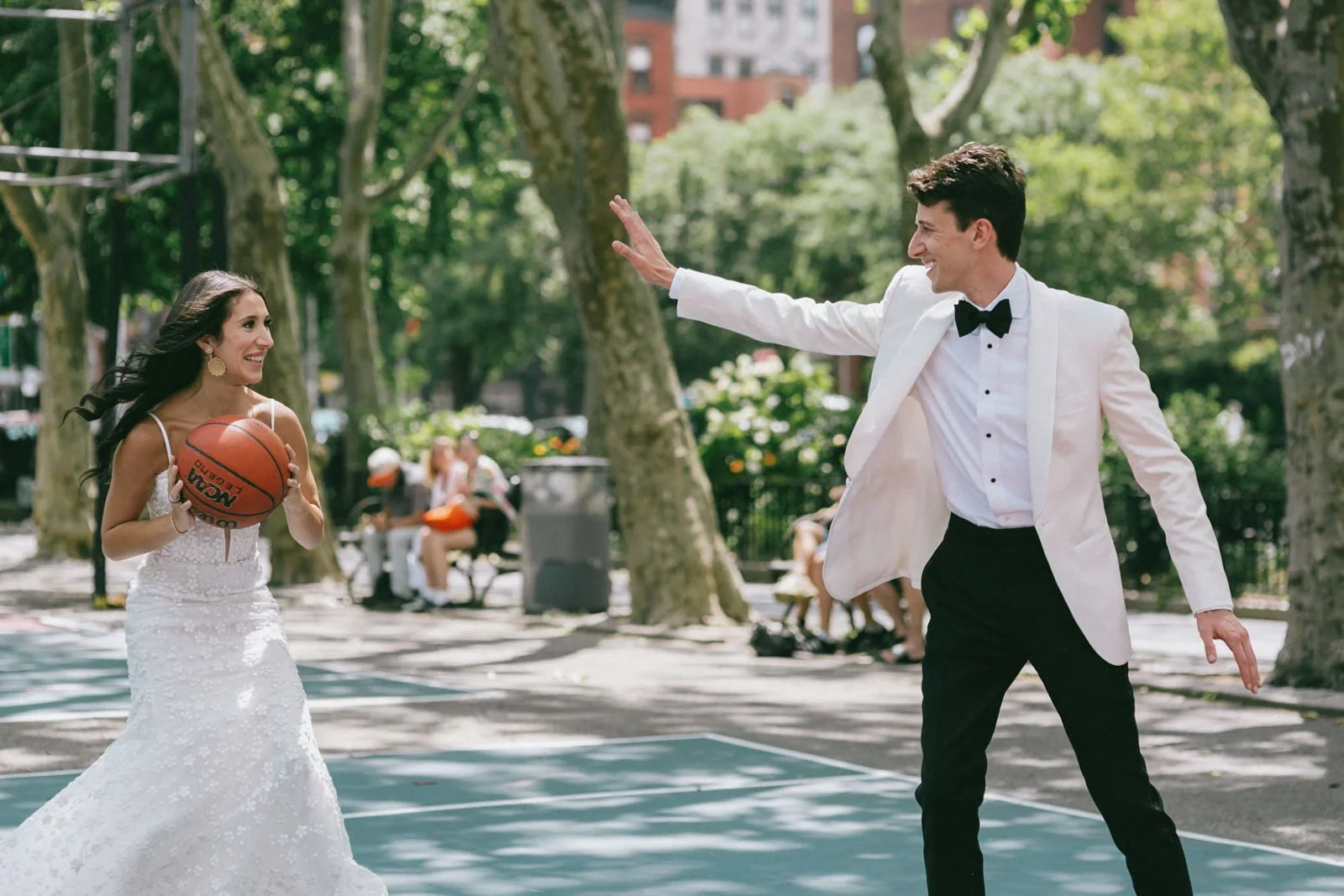A woman in a wedding dress holding a basketball walks towards a man in a tuxedo on an outdoor basketball court, smiling as he waves at her.